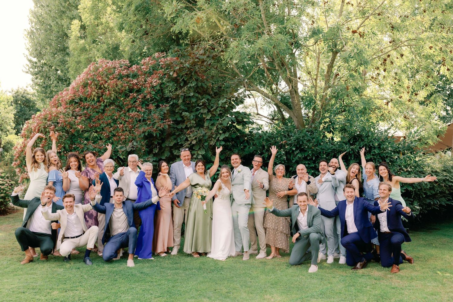 Large wedding party poses together on green lawn during outdoor summer celebration.