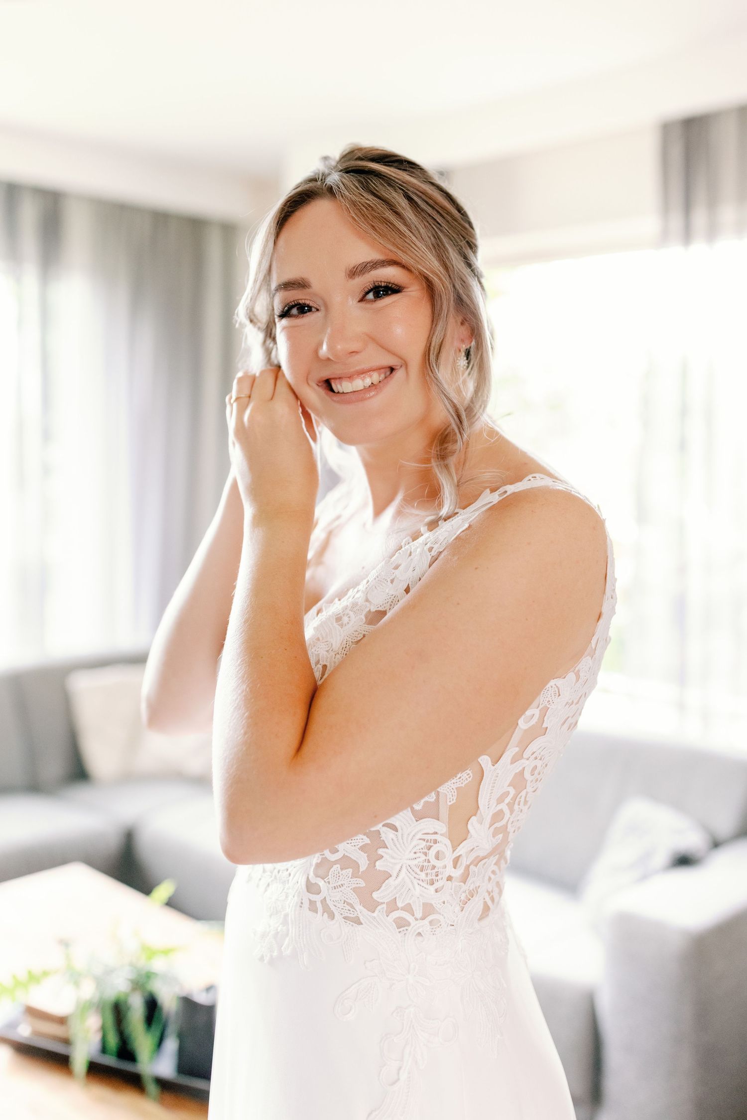 A bride in a white lace wedding dress smiles and adjusts her veil in a bright, modern room.