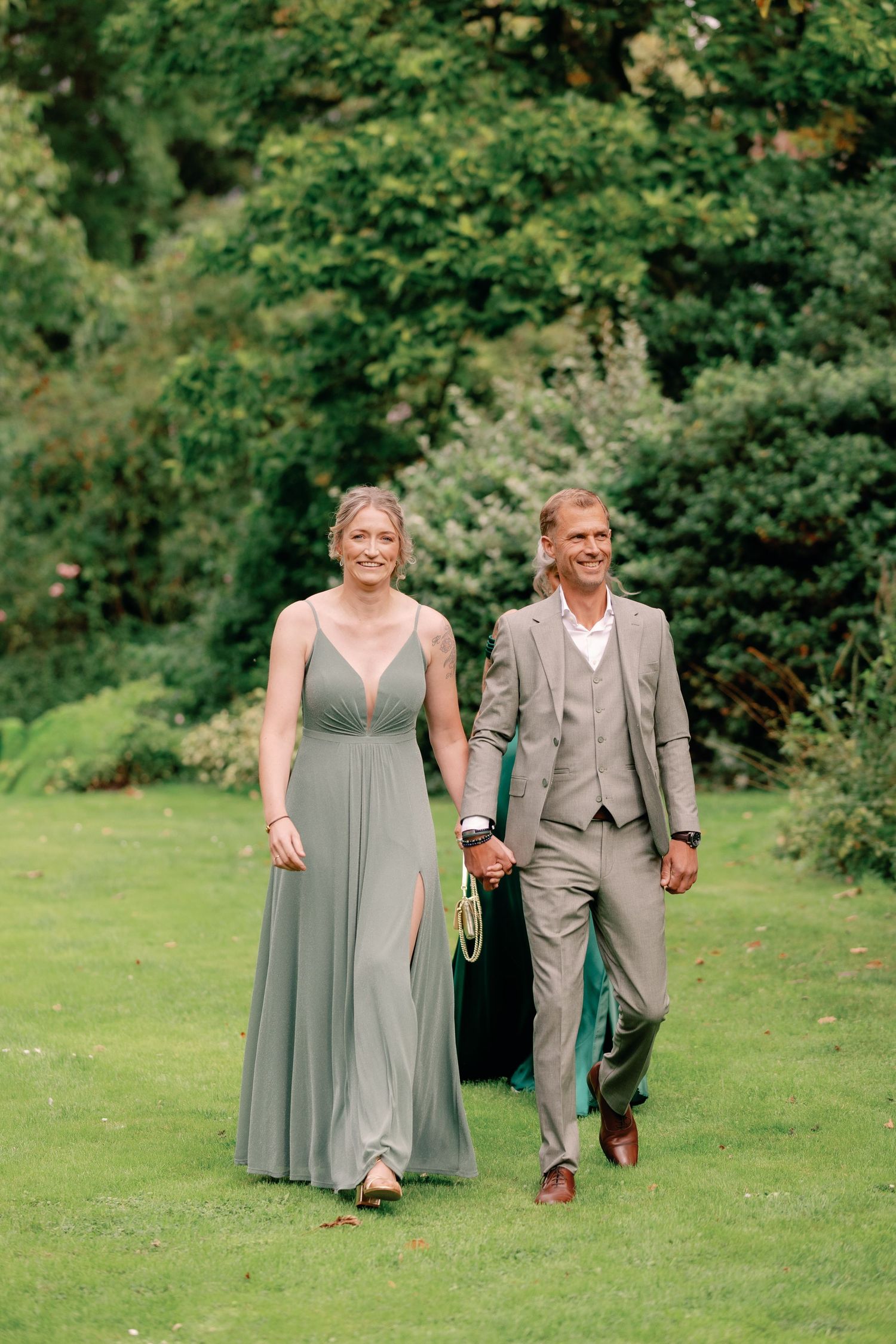 A couple in formal attire walks hand in hand across a lush green lawn surrounded by trees and bushes.