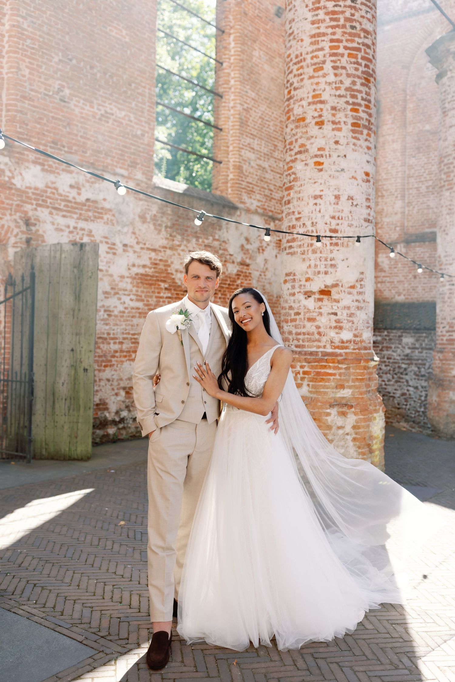 A couple in wedding attire poses against rustic brick columns with string lights and natural sunlight streaming in.