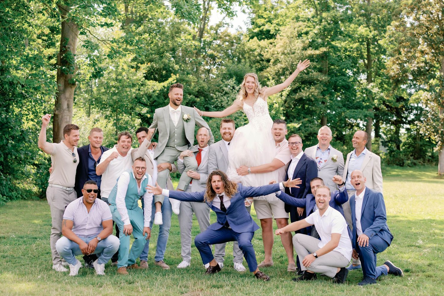 Fun wedding party poses playfully on green lawn with trees in background during summer outdoor celebration.