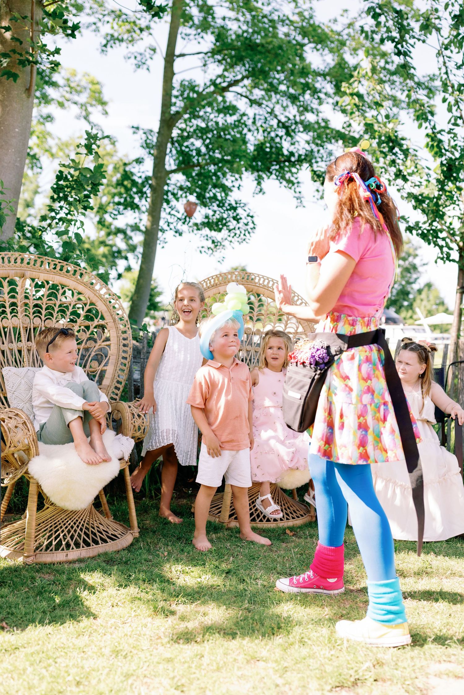 A group of children play together outdoors on a sunny day with peacock chairs and pink decorative elements.