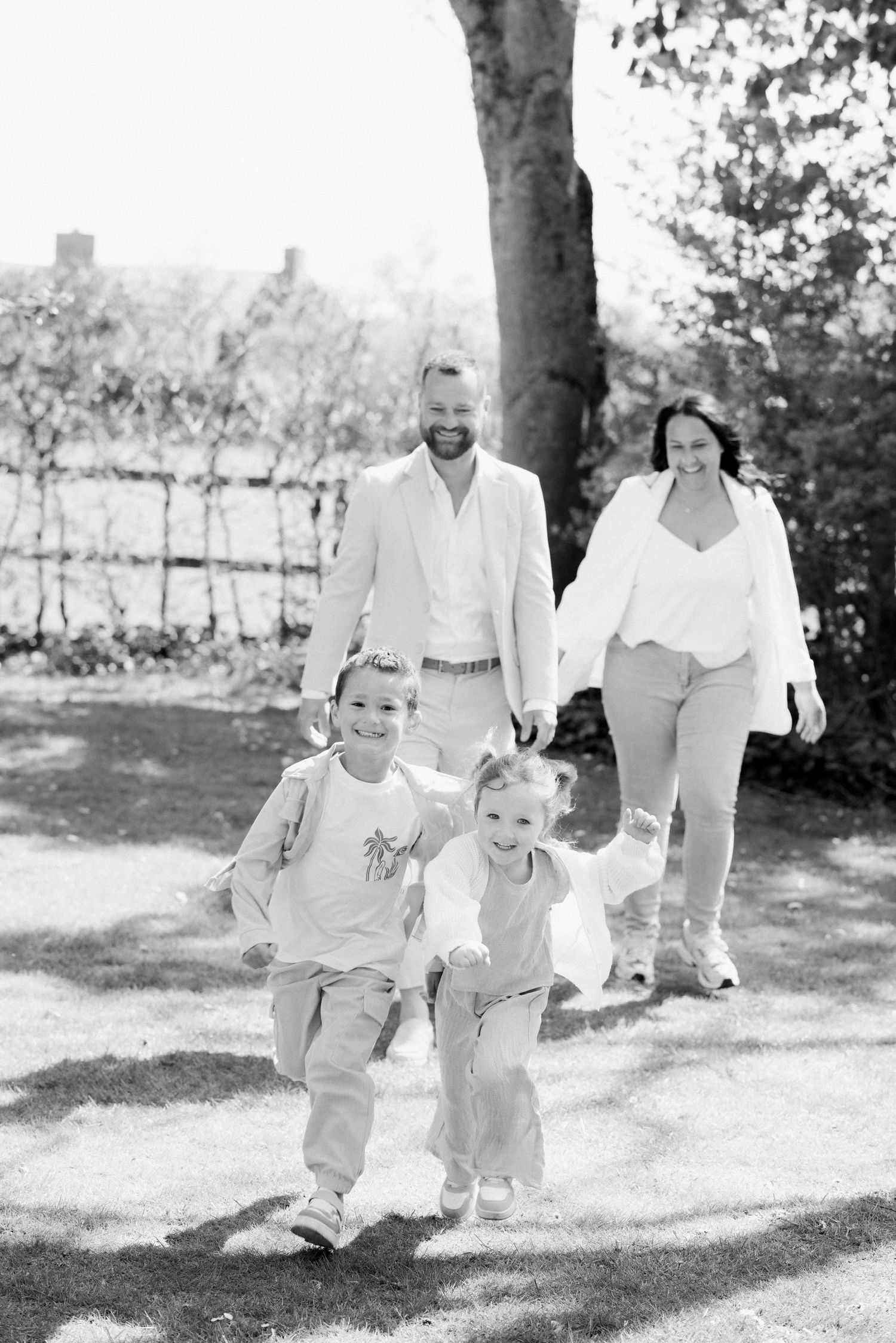 Black and white family photo of parents and two young children running outdoors in a sunny park setting.