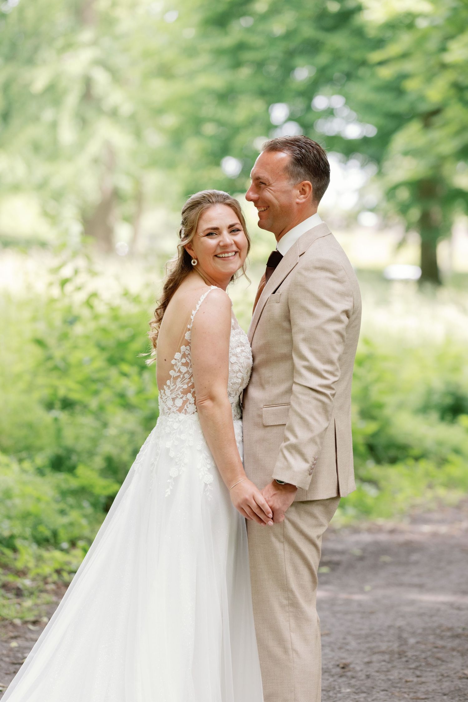 Smiling bride in backless lace wedding gown and groom in tan suit holding hands in a lush green park.