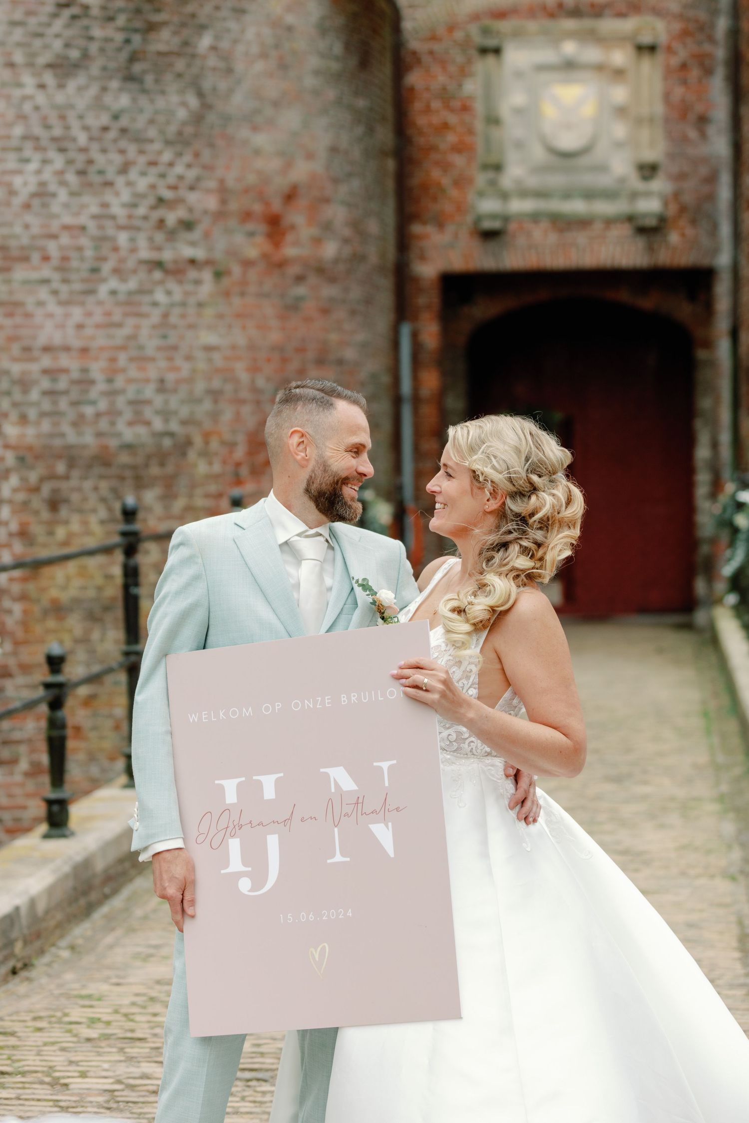 A newlywed couple shares a joyful moment while holding signs in front of a rustic brick wall.