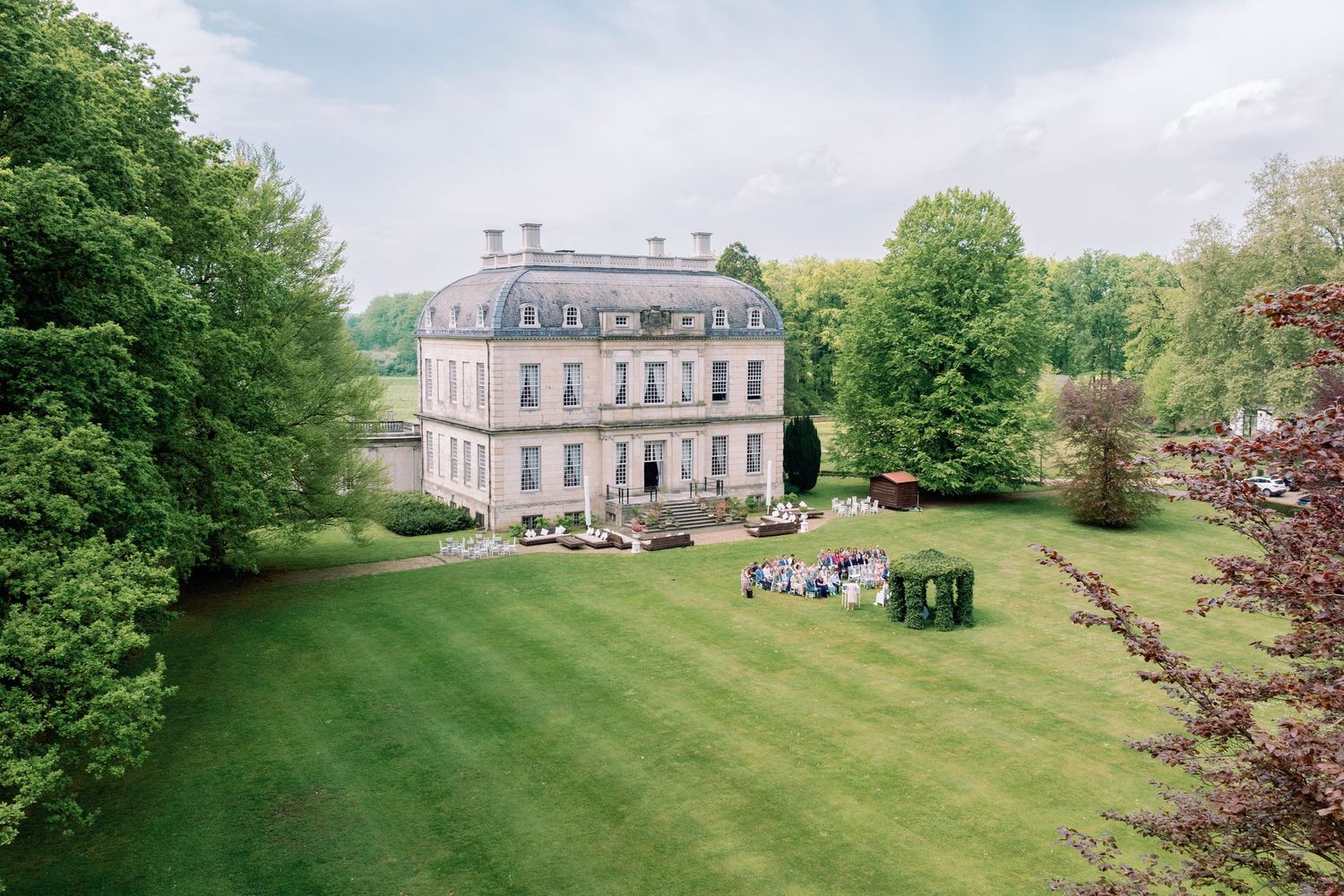 A panoramic view of a grand country estate with manicured lawns and classic architecture set among lush green trees.