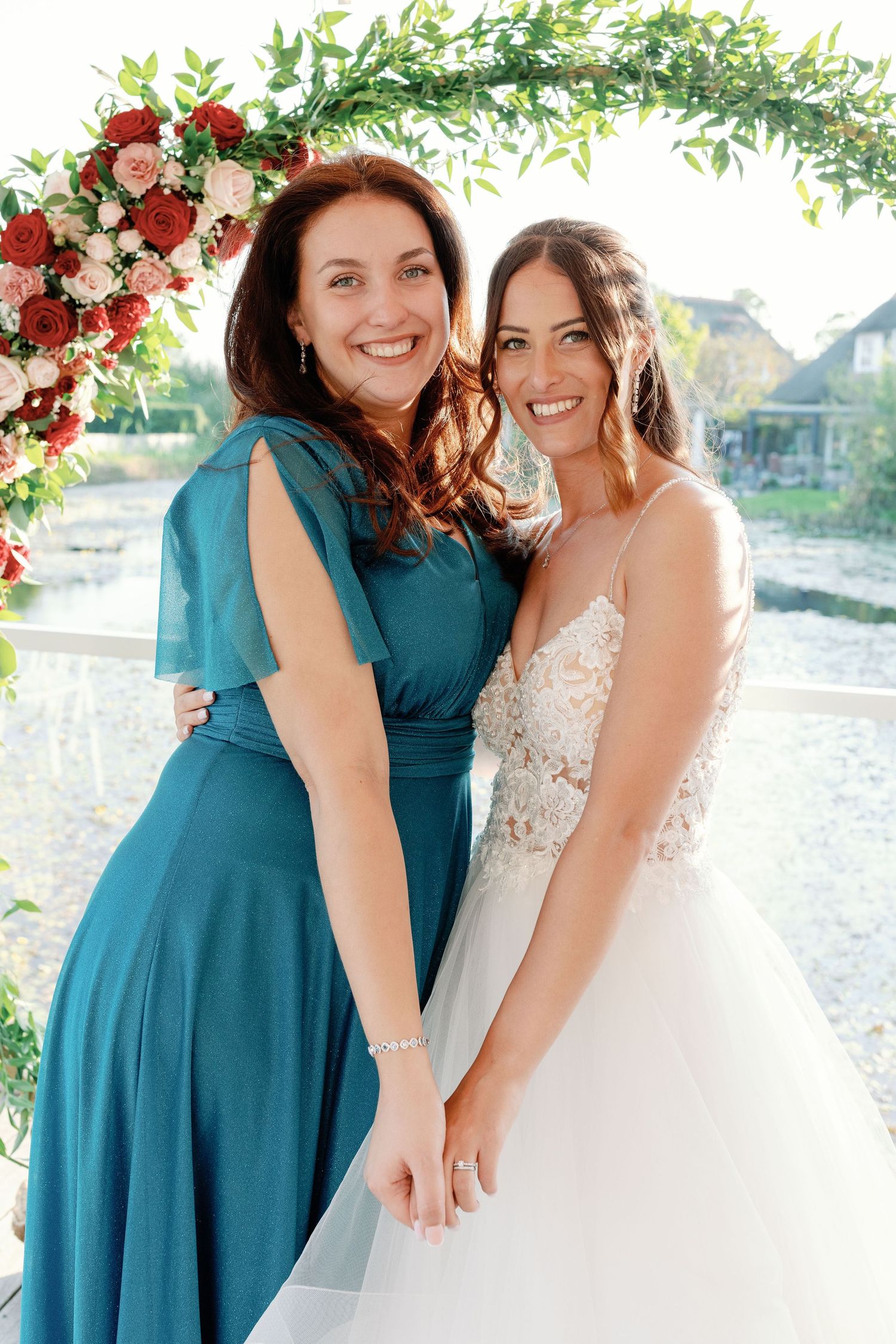 Bride and bridesmaid in teal dress share emotional moment under floral arch.