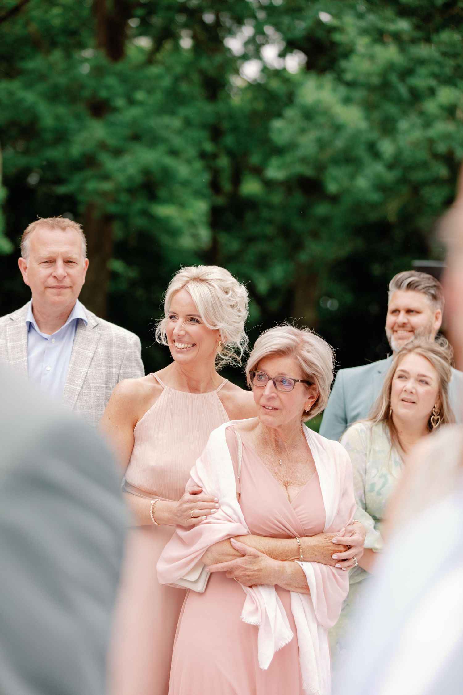 A group of people in light pink and white attire gather outdoors for a casual celebration in a garden setting.