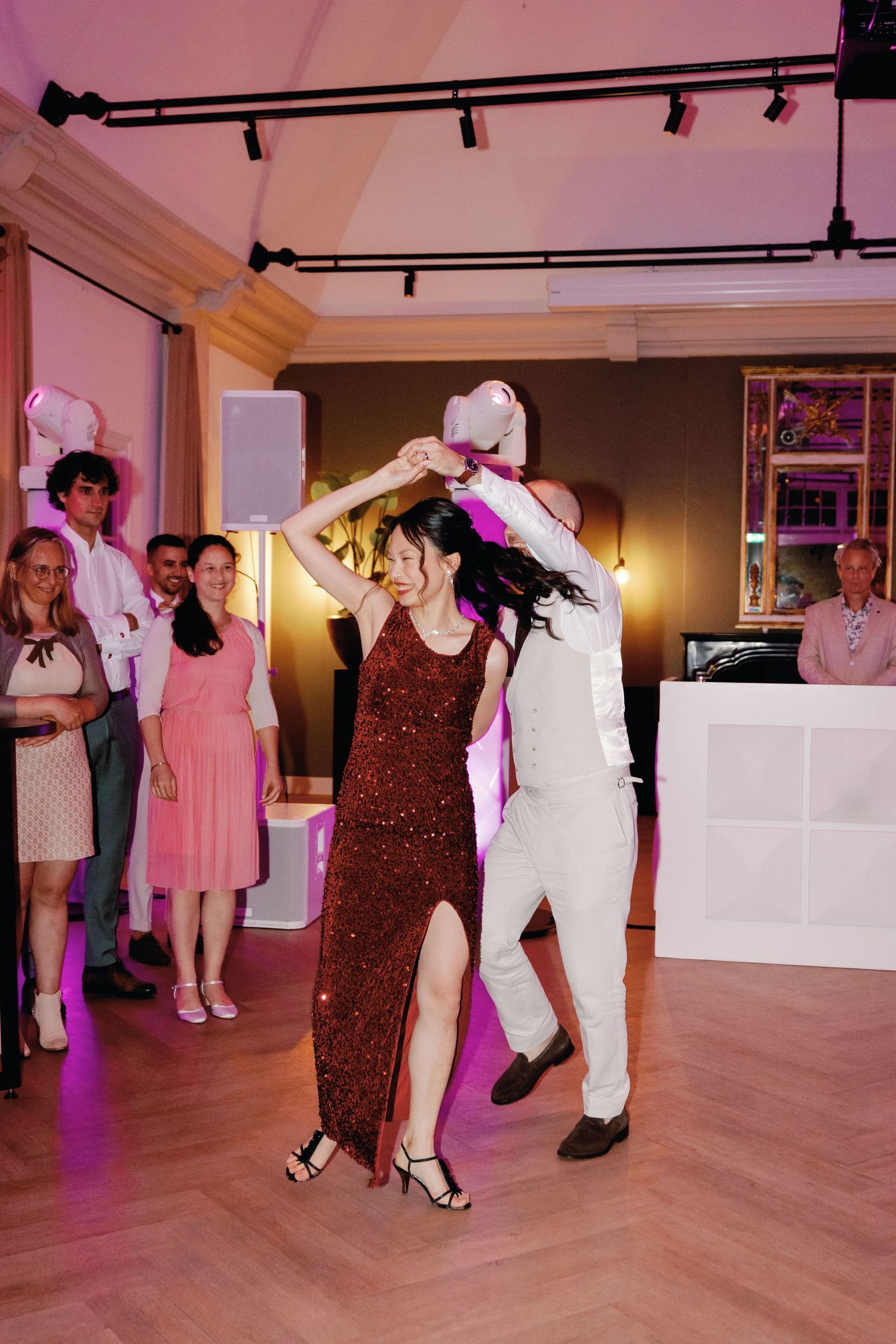 Couple dancing energetically at an indoor party venue with pink lighting and hardwood floors.