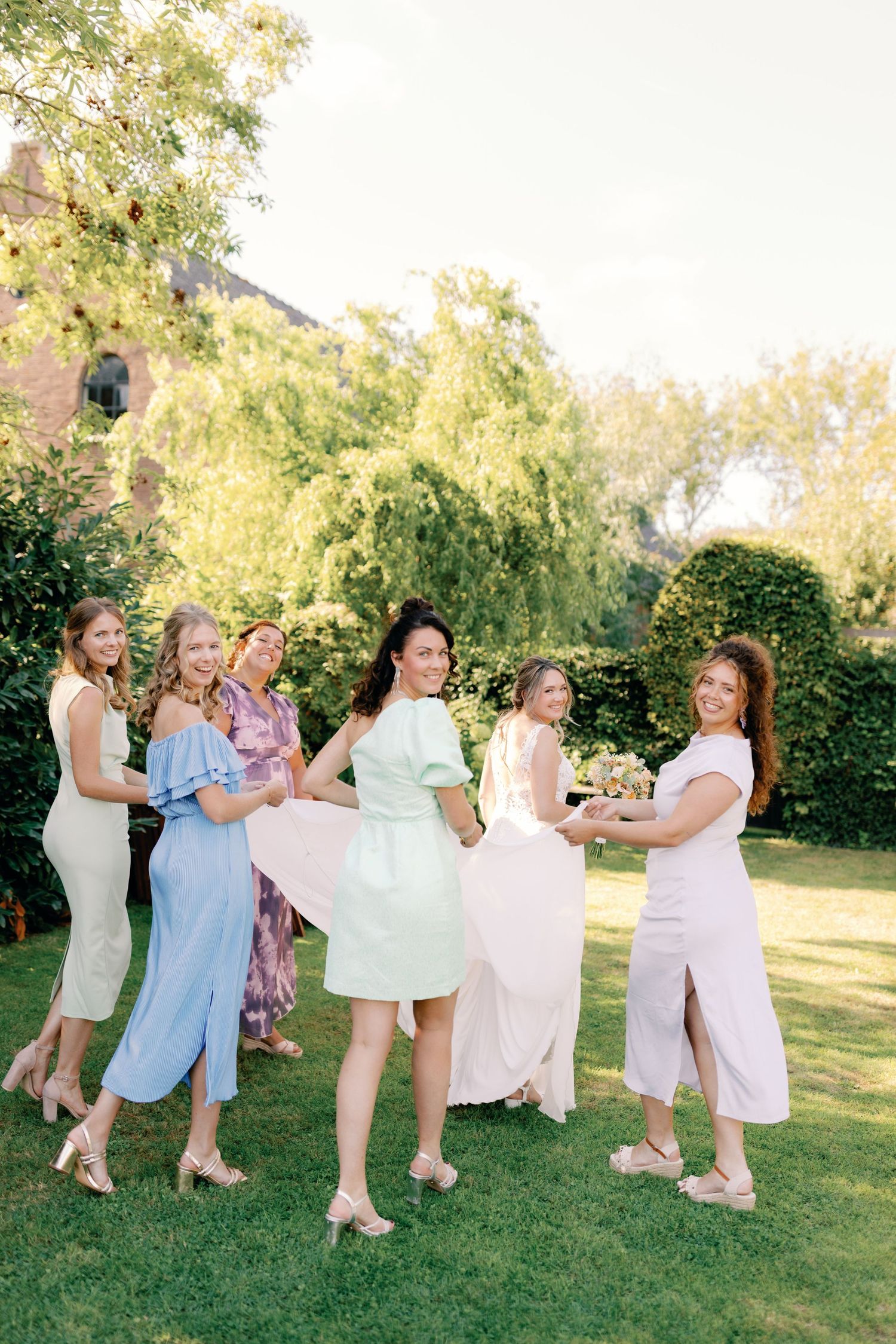 A group of bridesmaids in pastel dresses playfully dance and pose together on a sunny lawn during a wedding photoshoot.