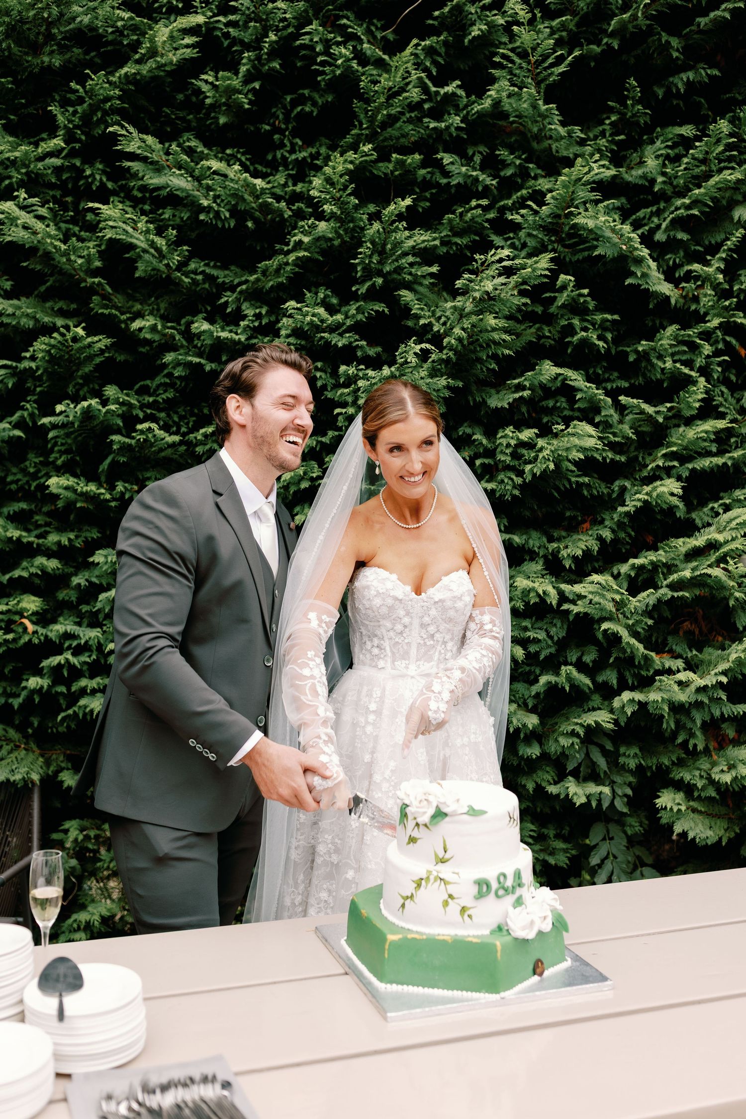 Newlywed couple shares a joyful moment while cutting their green and white wedding cake together in a garden setting.