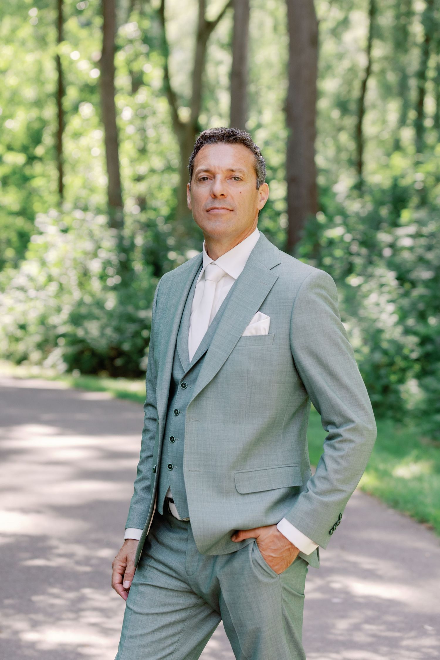 A person in a light gray three-piece suit poses confidently on a tree-lined path in a natural outdoor setting.
