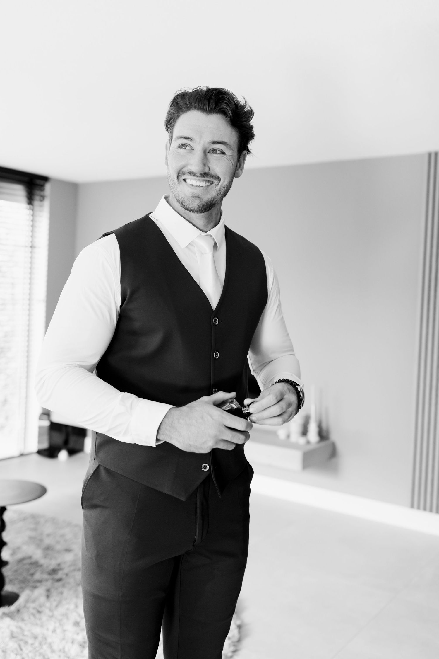 A person in formal attire adjusts their black vest while smiling in a bright white room.