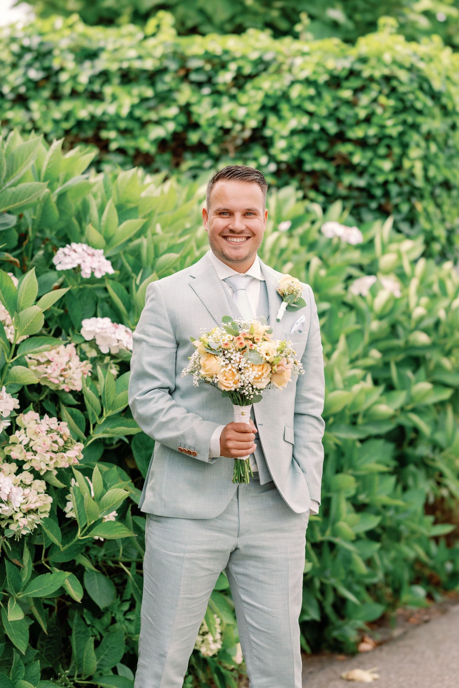 A light gray suited figure holds a peach and cream colored wedding bouquet against a lush garden backdrop.