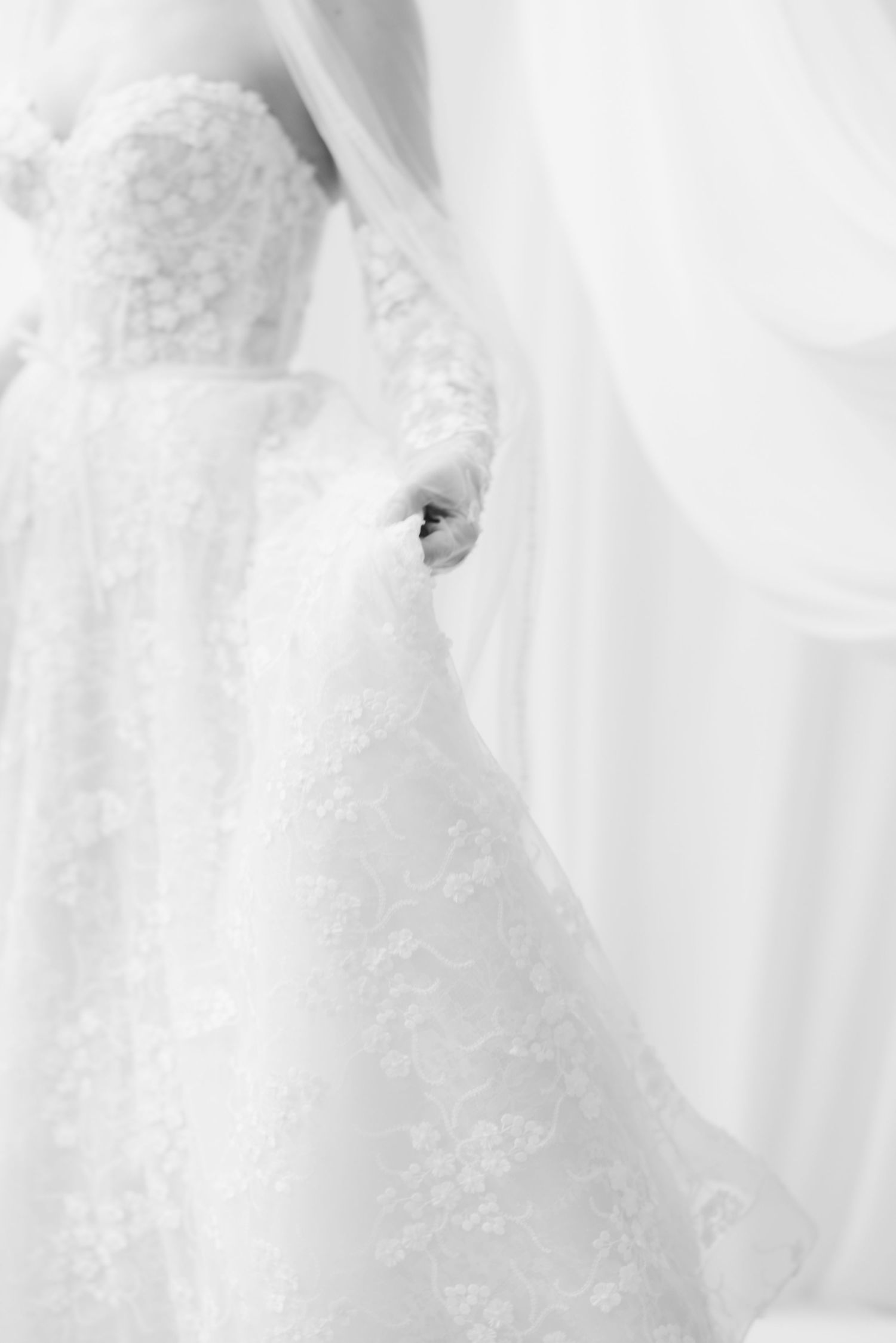 Black and white photo of a bride in a floral lace wedding gown with sheer sleeves holding the skirt beside soft draped curtains.