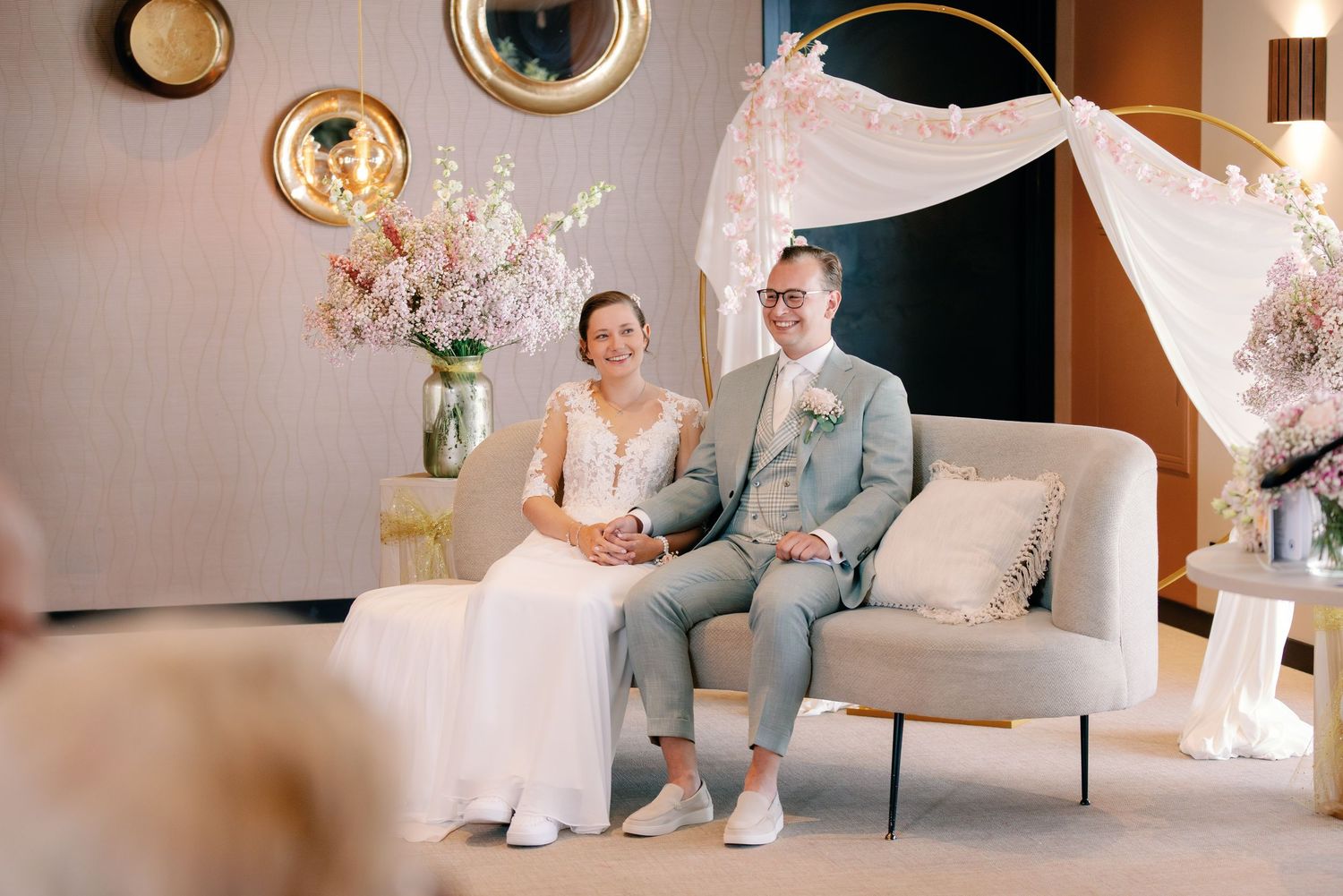 Bride and groom seated on elegant sofa during intimate wedding ceremony with soft pink floral decorations.