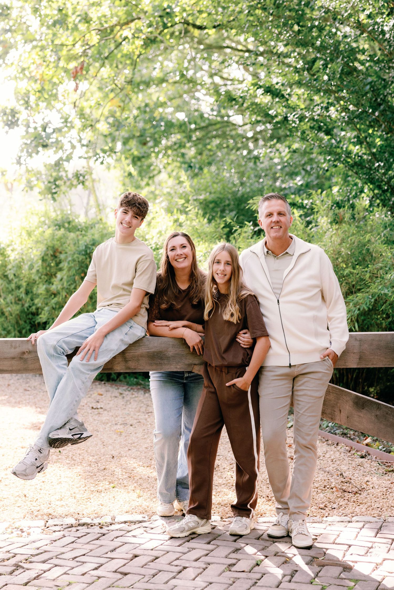 Family of four posing outdoors by a wooden fence, with a teen boy sitting on the rail and a girl, woman and man standing on a brick path.