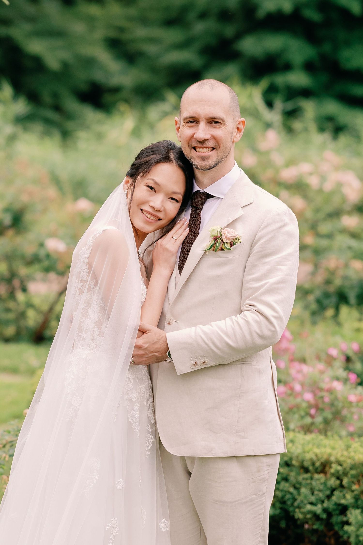 A happy couple embraces outdoors during their wedding ceremony surrounded by pink flowers and greenery.
