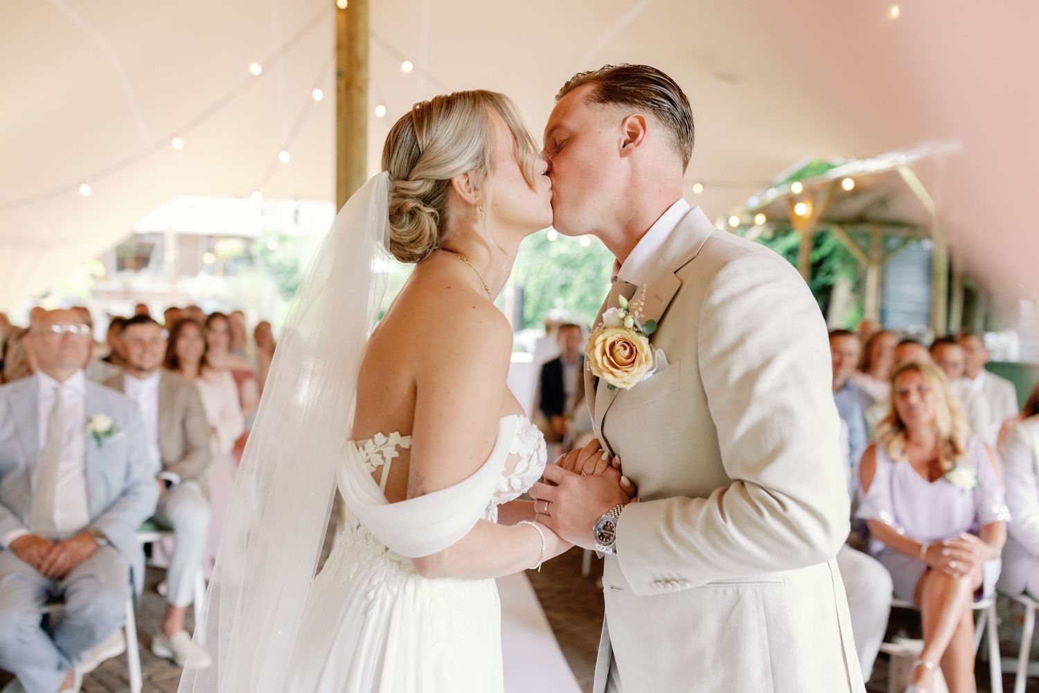 A couple shares their first kiss as newlyweds during a wedding ceremony with guests watching in the background.