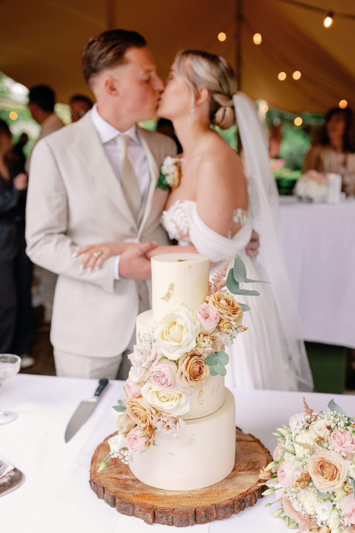 Elegant white wedding cake decorated with peach roses sits on rustic wooden slice at reception.