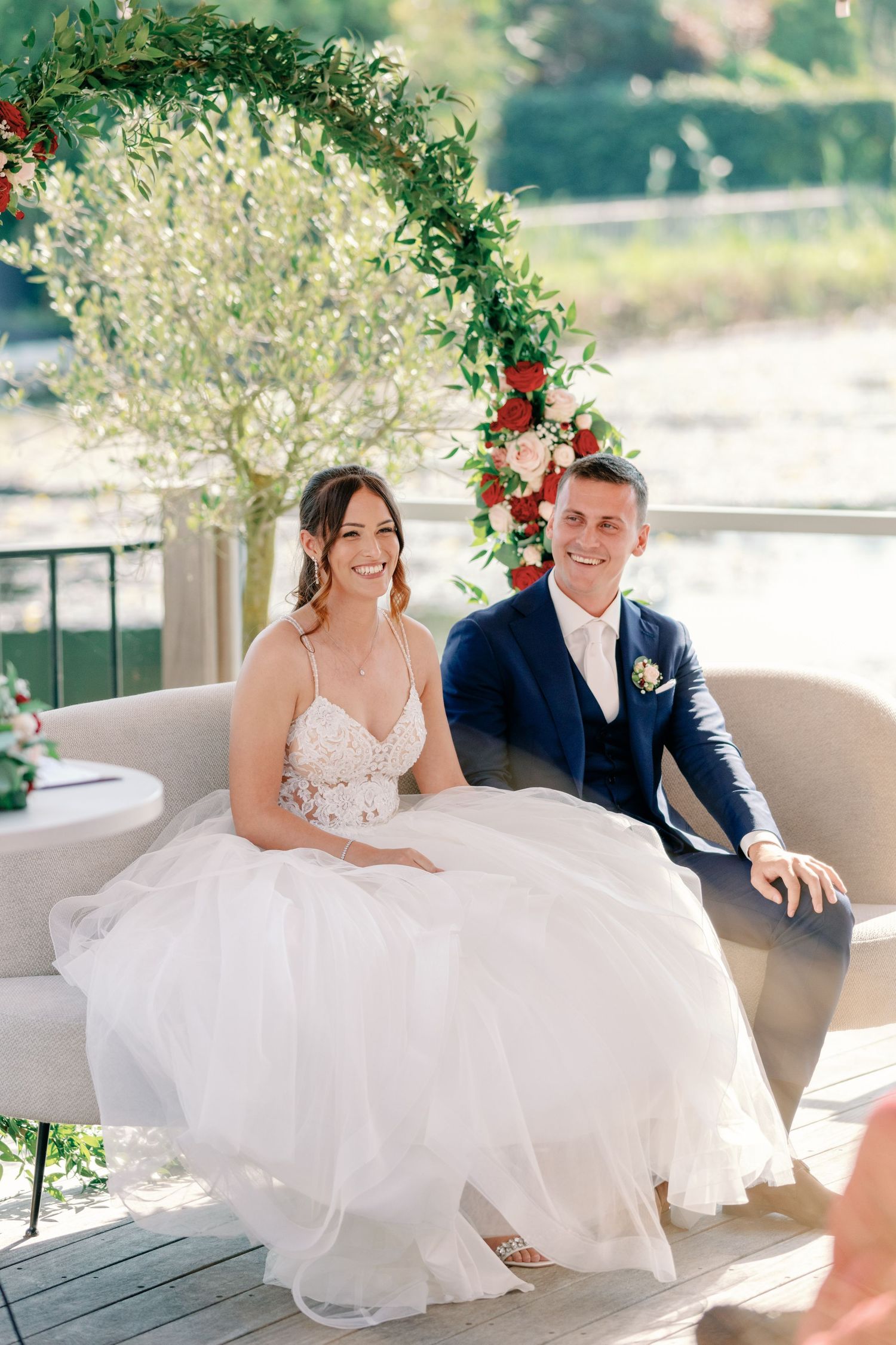 Newlyweds sit together on an outdoor couch decorated with floral arrangements and greenery.