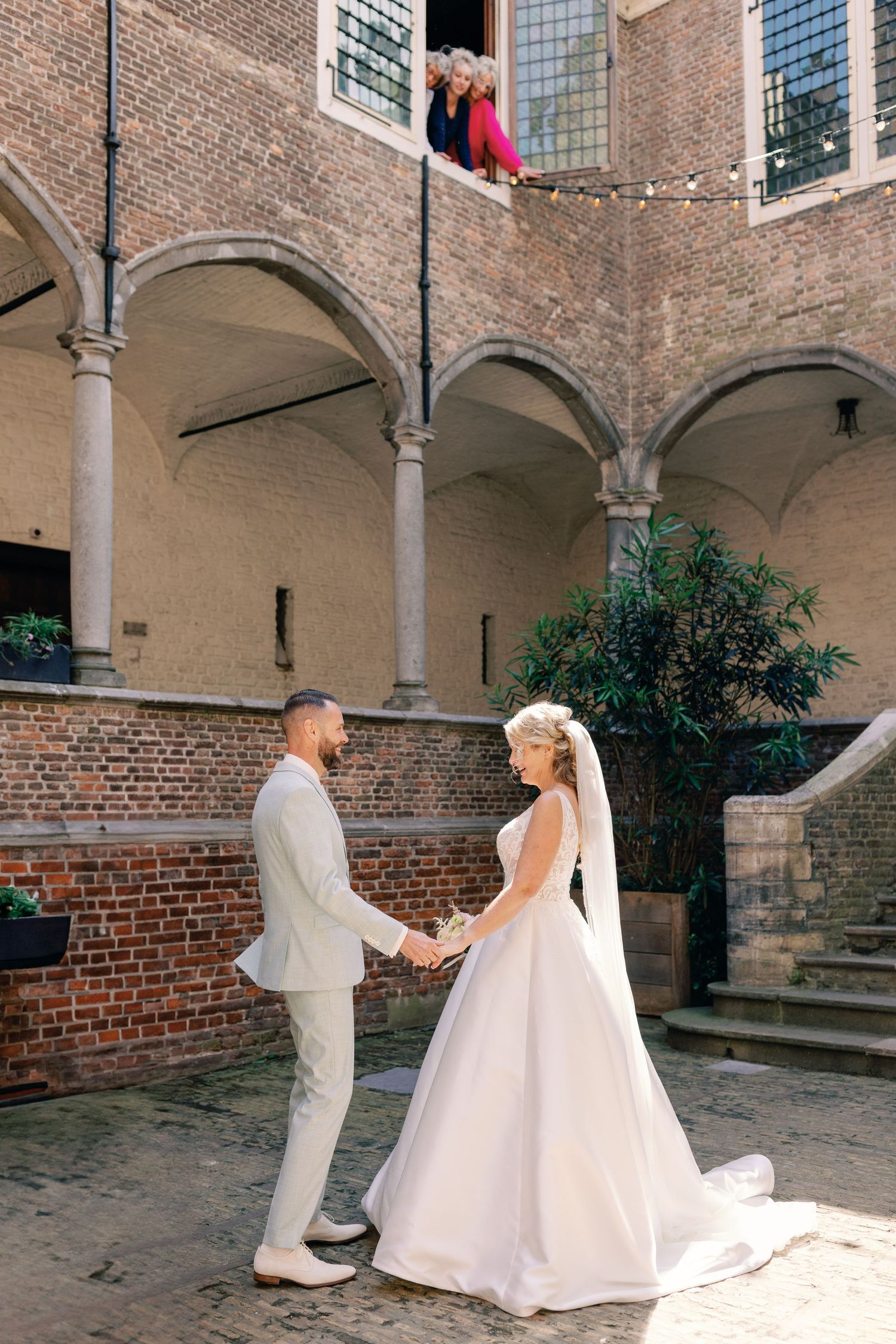 A couple shares a first look moment in a brick courtyard with arched columns and string lights overhead.