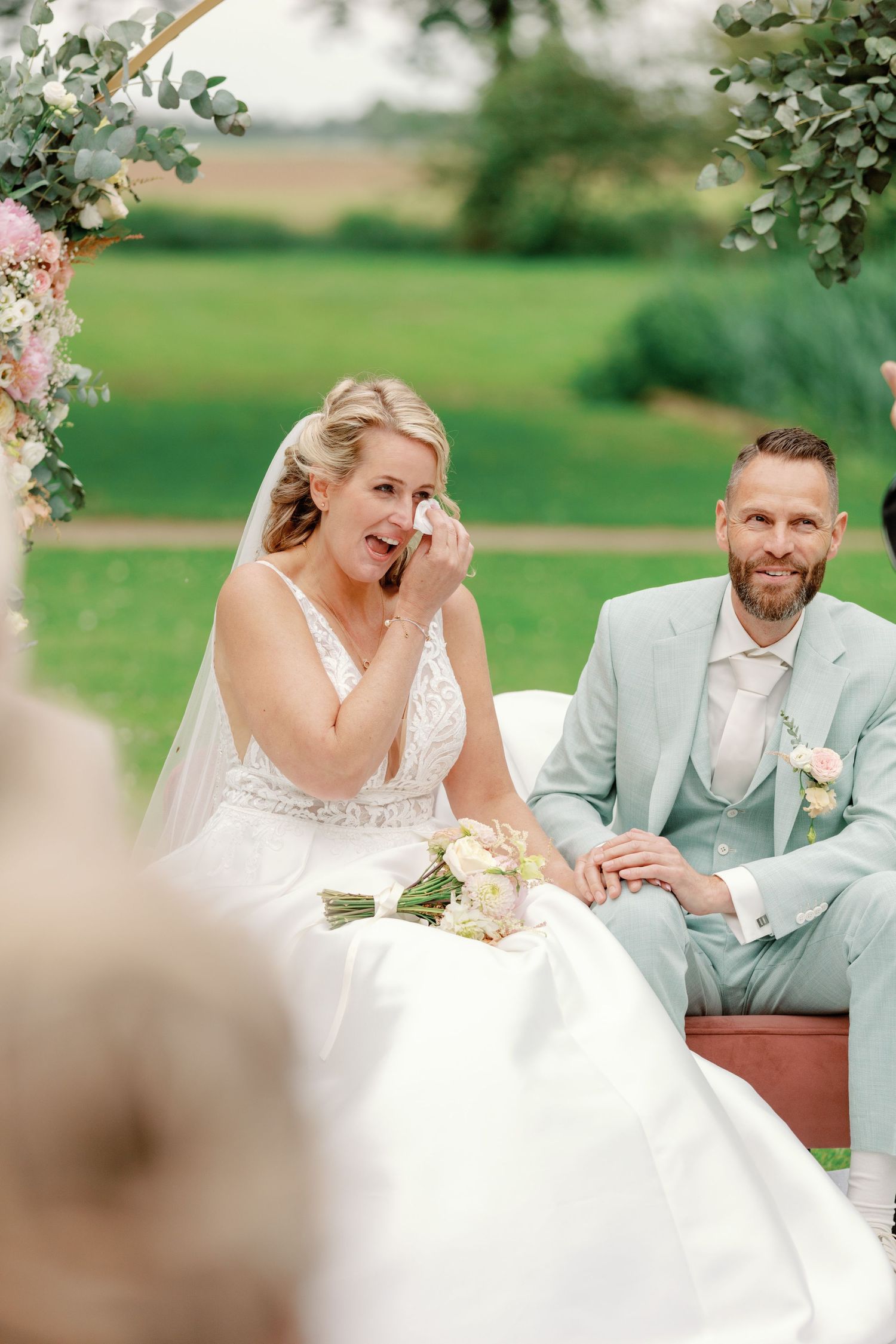 A couple shares a joyful moment together on a bench during their outdoor wedding photoshoot.