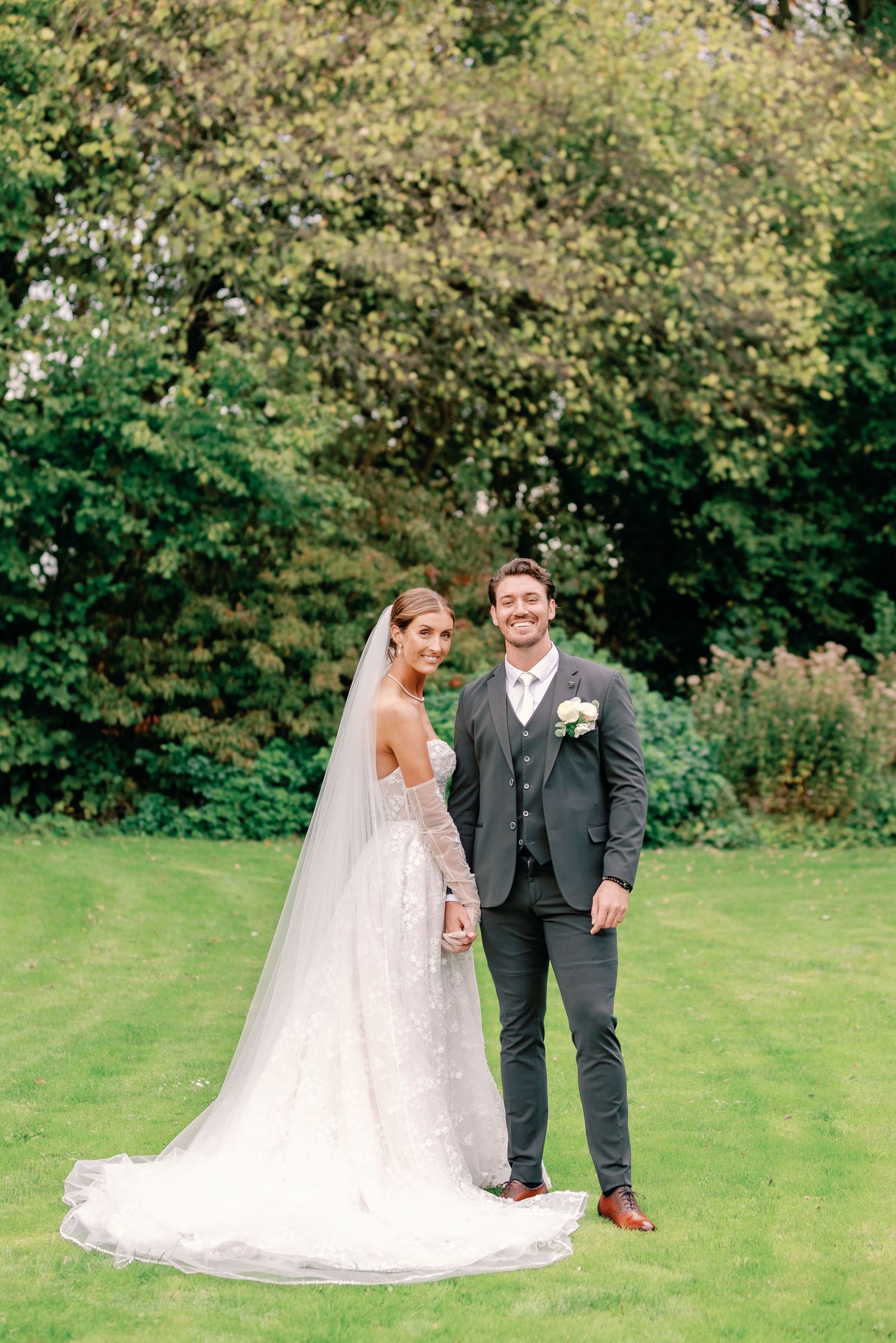 A couple in wedding attire poses on a lush green lawn with autumn trees in the background.