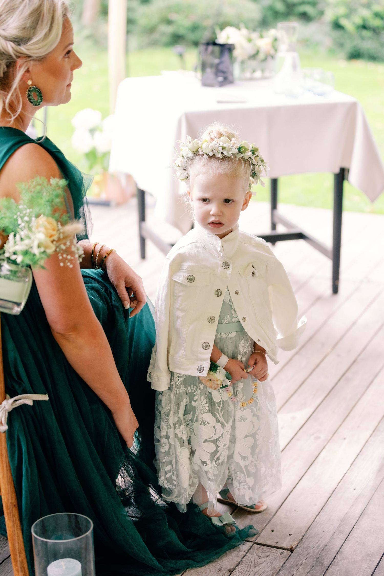 A baby in white formal wear sits with someone wearing an emerald green dress on a wooden deck during a special occasion.