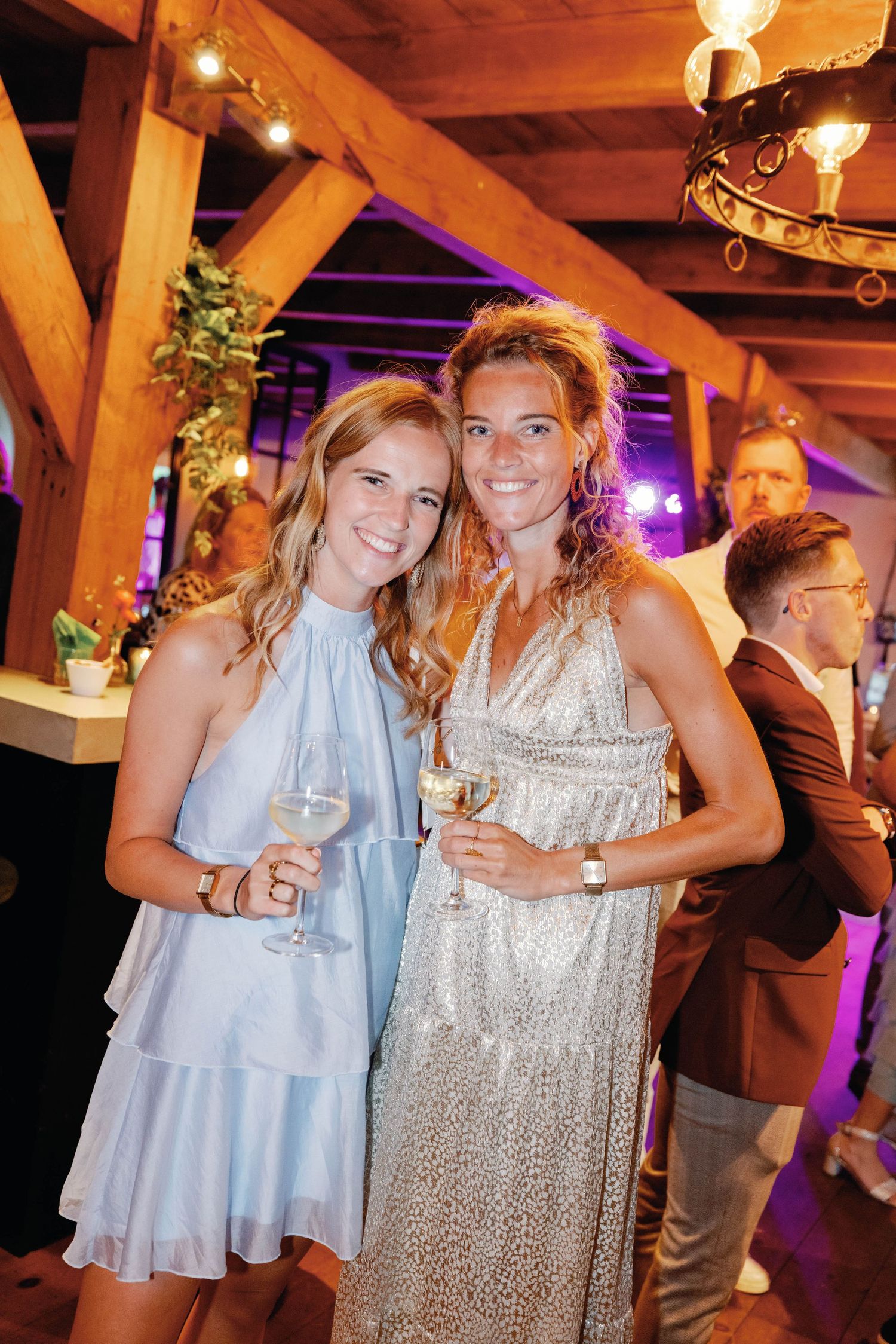 Two women in party attire smile and pose together while holding wine glasses at an indoor event with wooden beams.