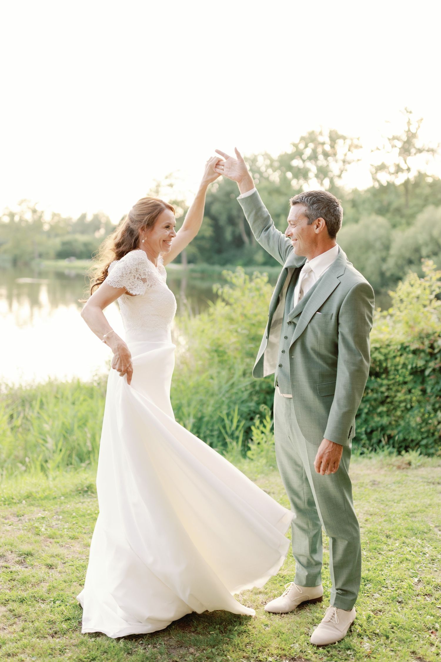 A couple dances together outdoors in wedding attire as sunlight filters through trees in the background.