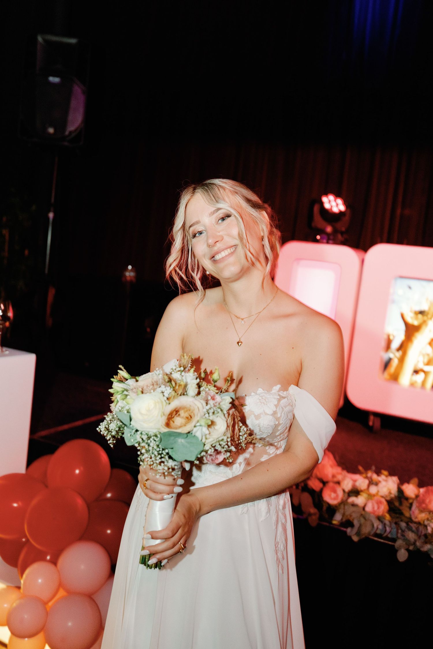 A bride in a white wedding dress holds a floral bouquet at a dimly lit reception with pink balloons and lighting.