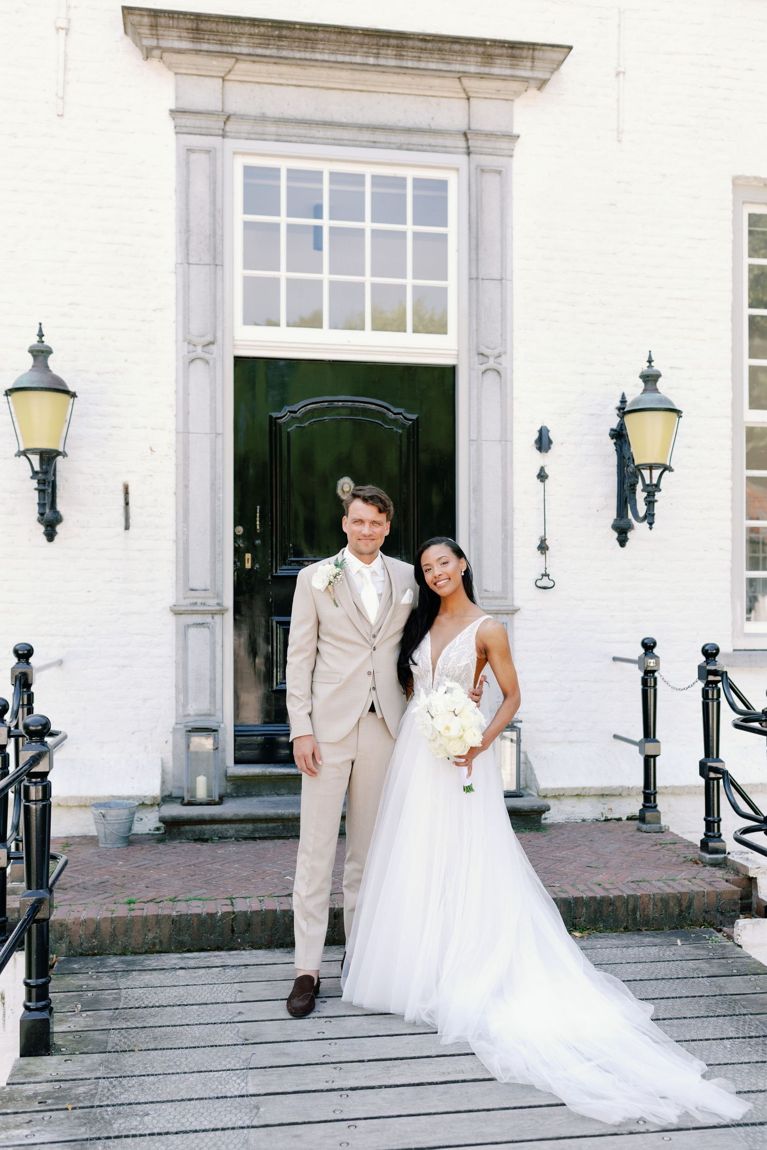 A wedding couple stands in front of a classic white building entrance with green door and lantern sconces.