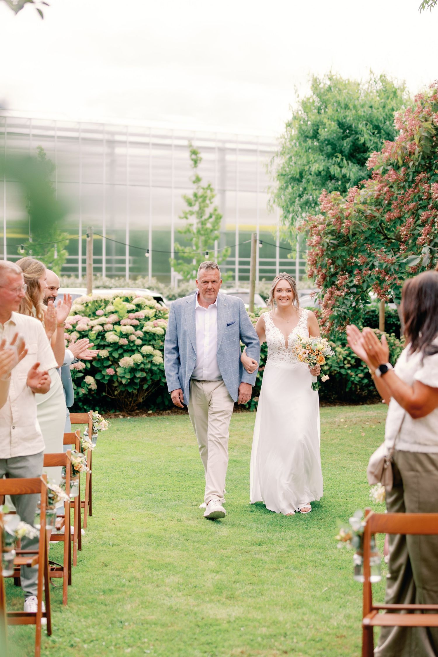 Wedding couple walks down the aisle between wooden chairs on a lush green lawn with flowering bushes in the background.