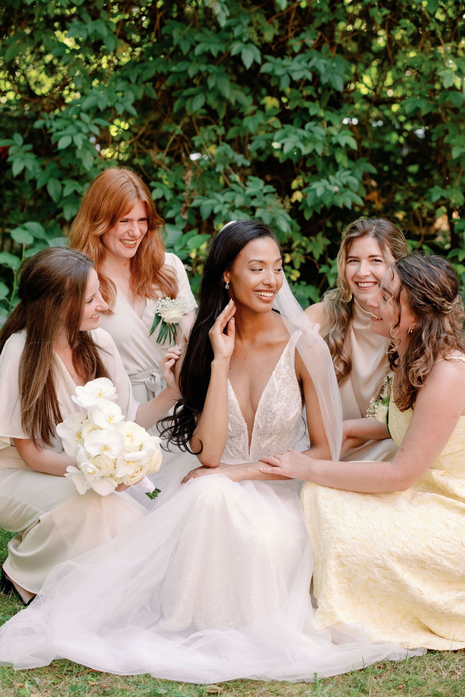 Bridesmaids in light colored dresses gather around bride in white gown outdoors with greenery backdrop.