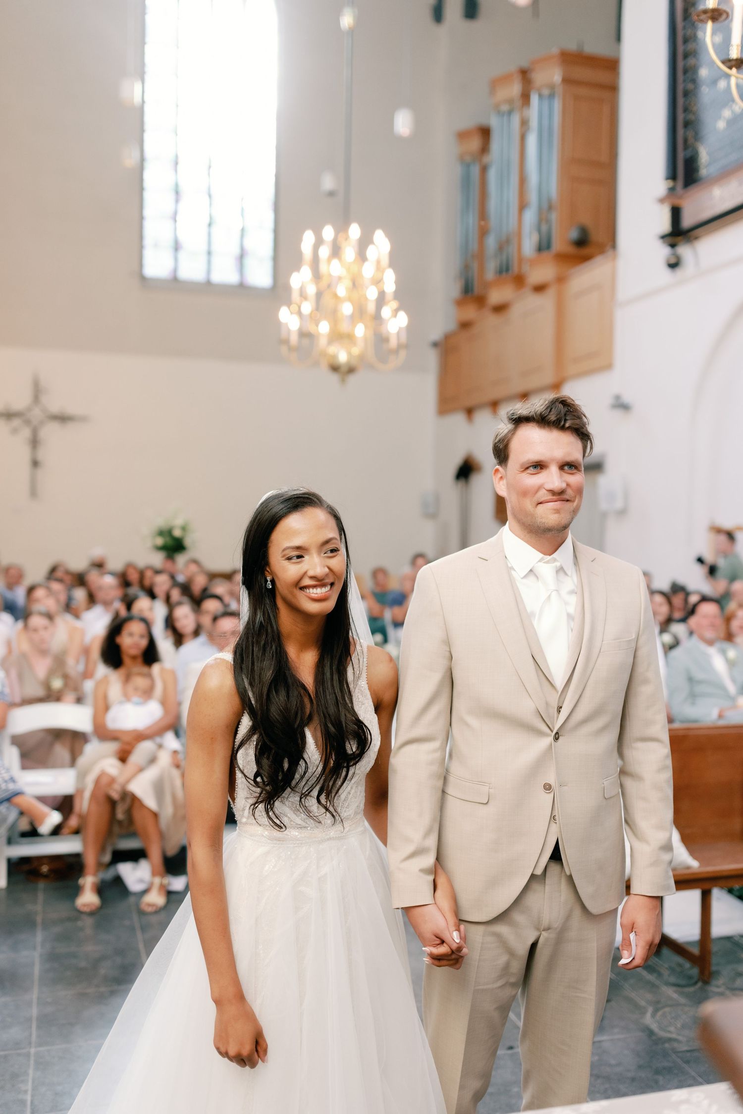 A bride and groom share joyful moments inside a bright church with elegant chandeliers and white decor.