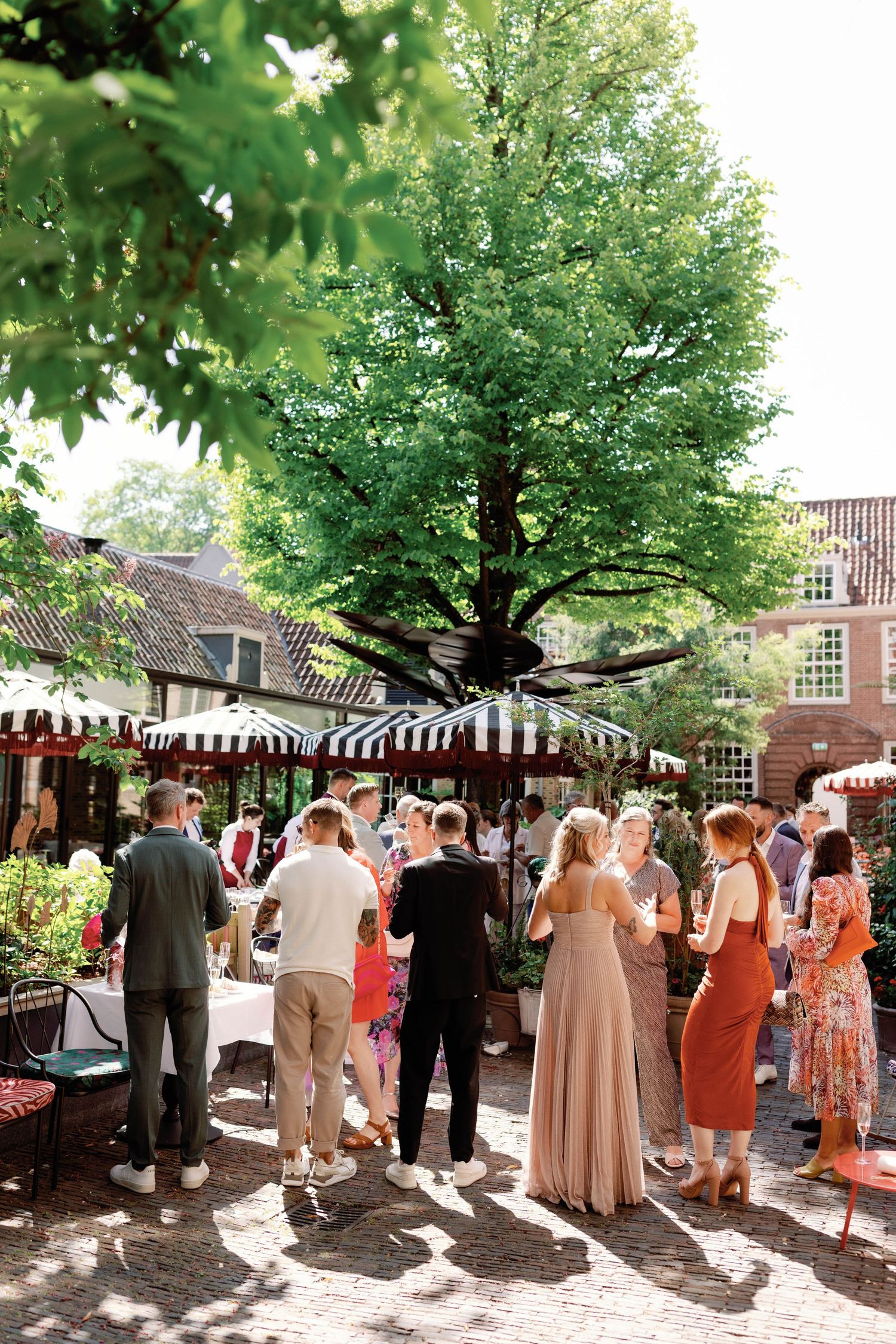 People enjoying a sunny outdoor market with striped umbrellas and vendors beneath lush green trees in an urban setting.