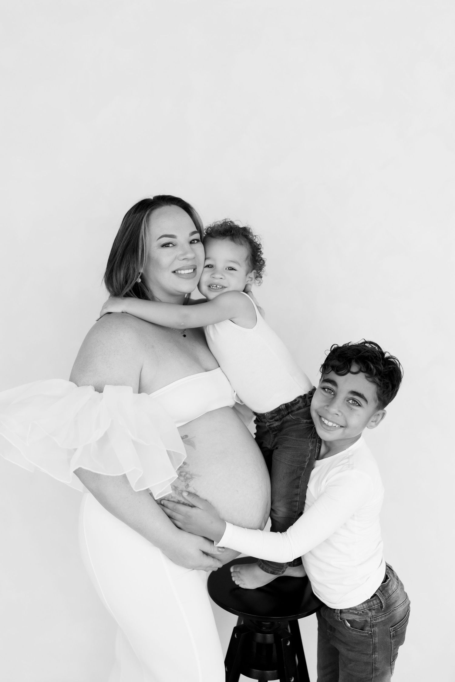 A black and white maternity photo showing a pregnant woman in white dress embracing a child and partner against light background.