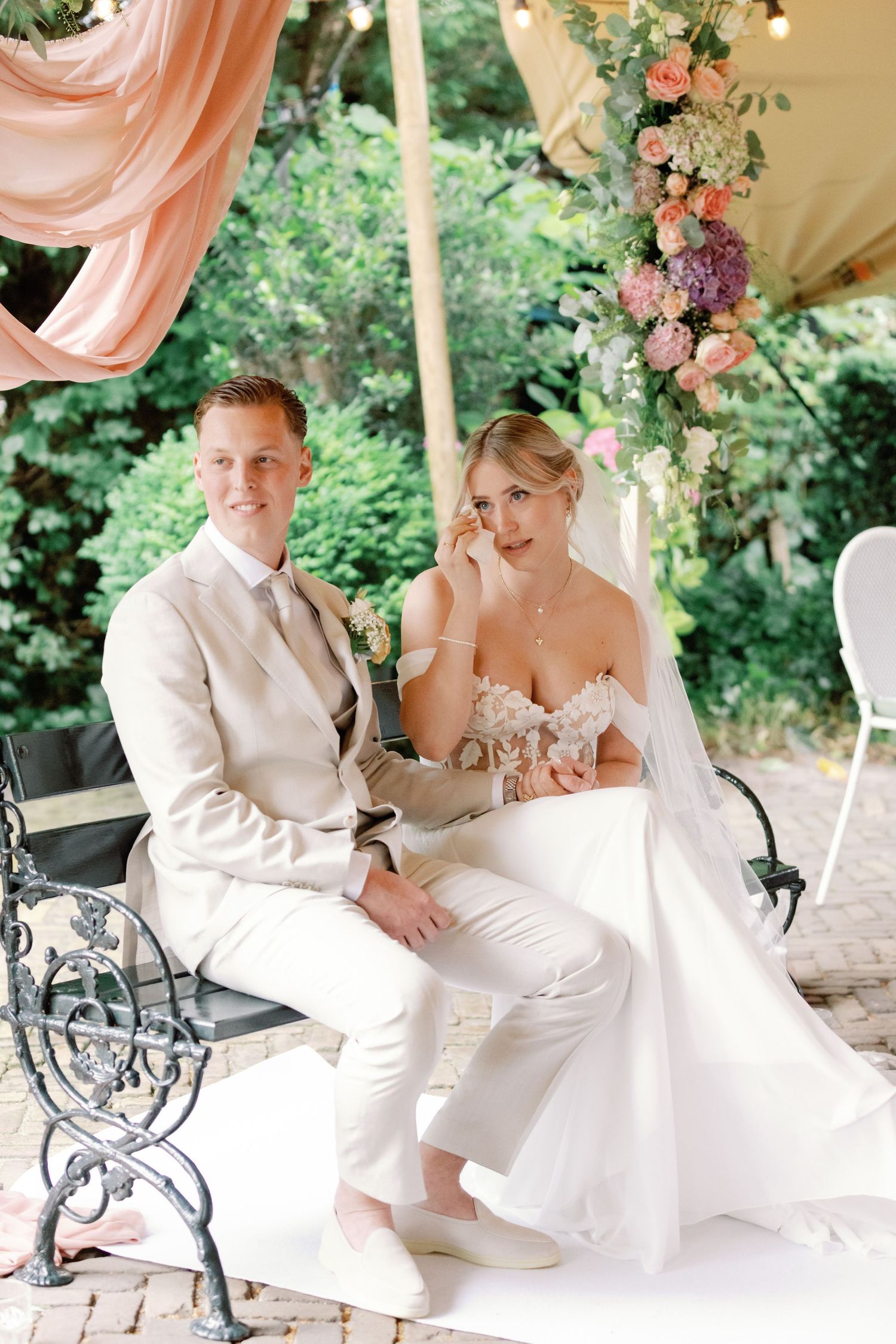 A romantic wedding couple shares intimate moments on a garden bench under pink drapery and string lights.