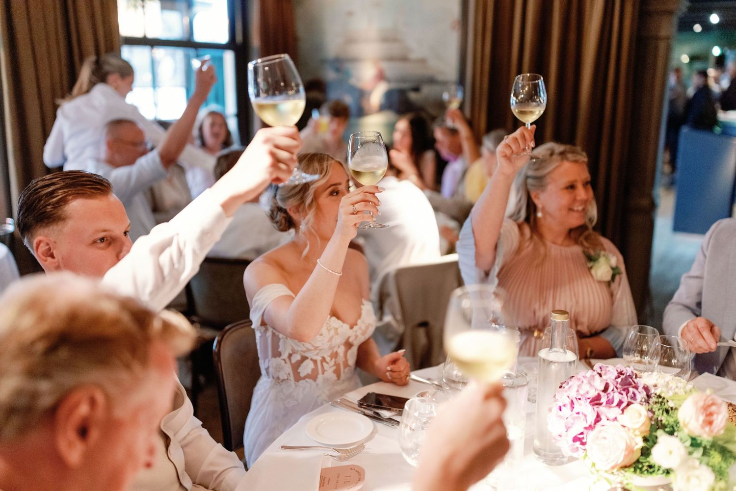 Wedding reception guests raise their wine glasses in a celebratory toast at an elegant indoor venue.