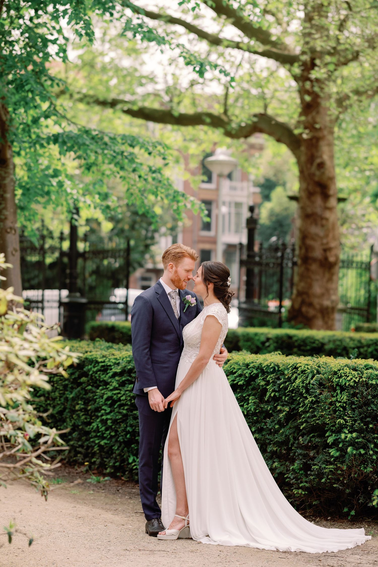 A couple in wedding attire share an intimate moment in a lush garden setting with manicured hedges and trees.