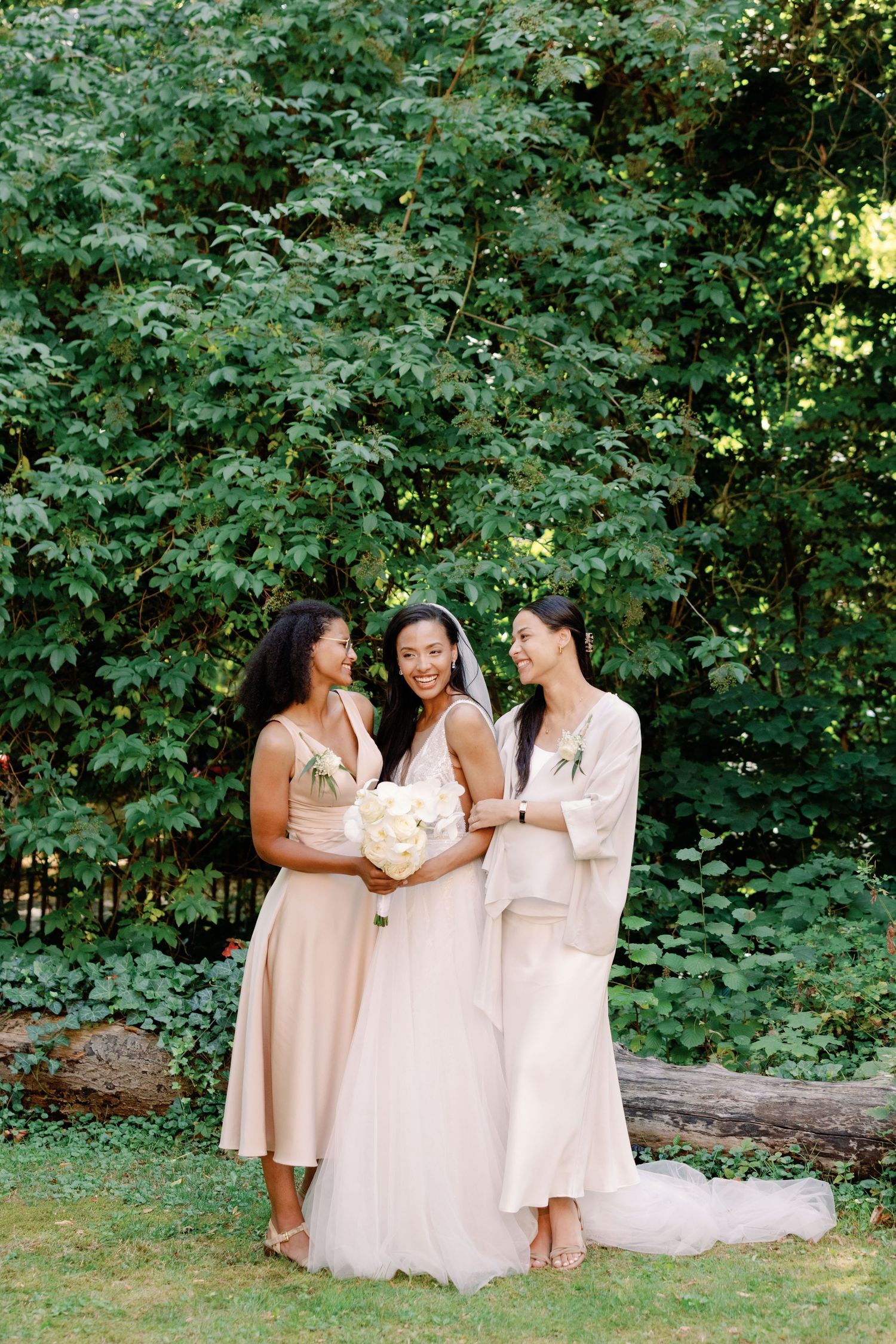 Three women in blush-toned dresses share a joyful moment together in a lush garden setting during a wedding celebration.