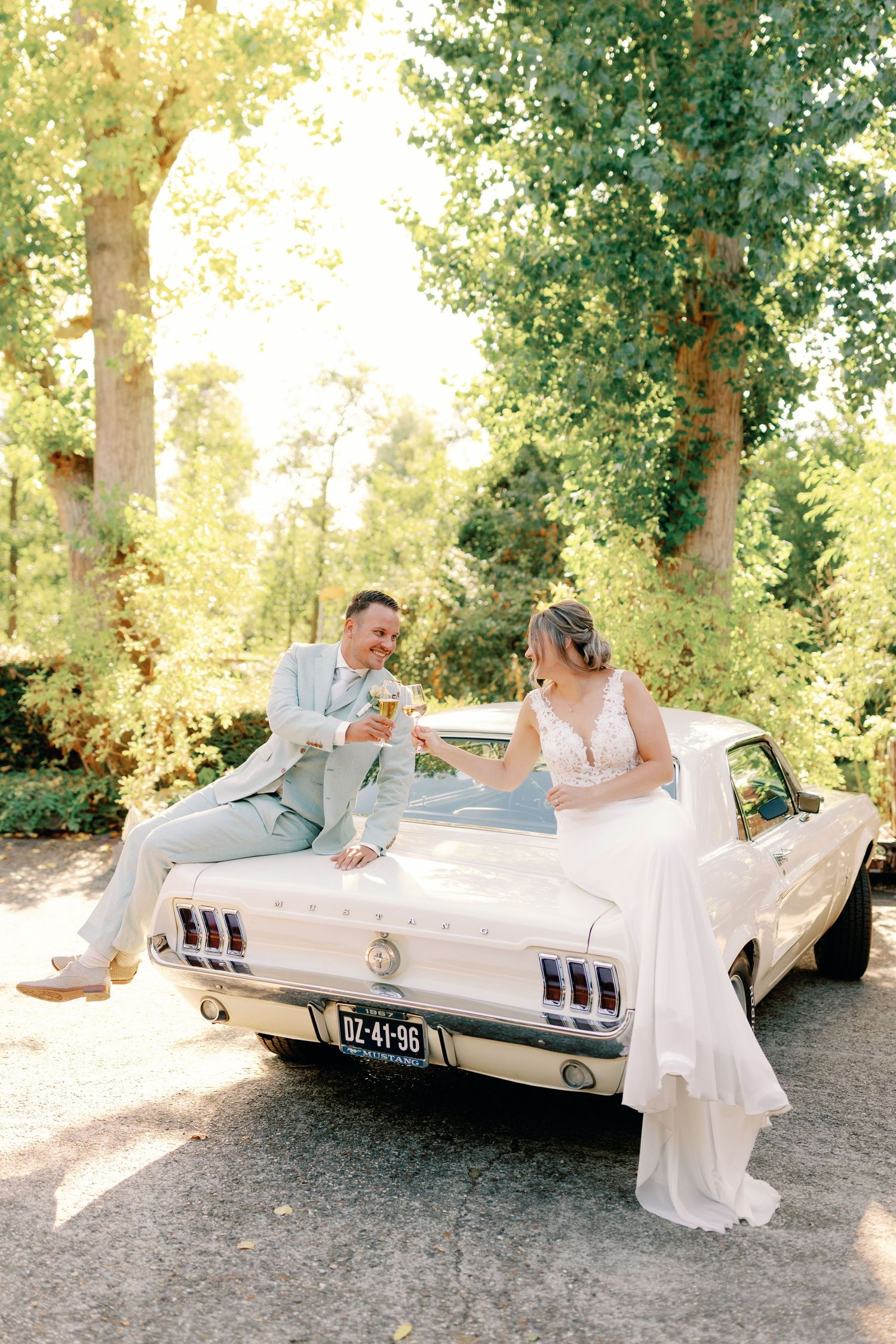 A couple in wedding attire poses playfully by a classic white vintage car in a sunlit outdoor setting.