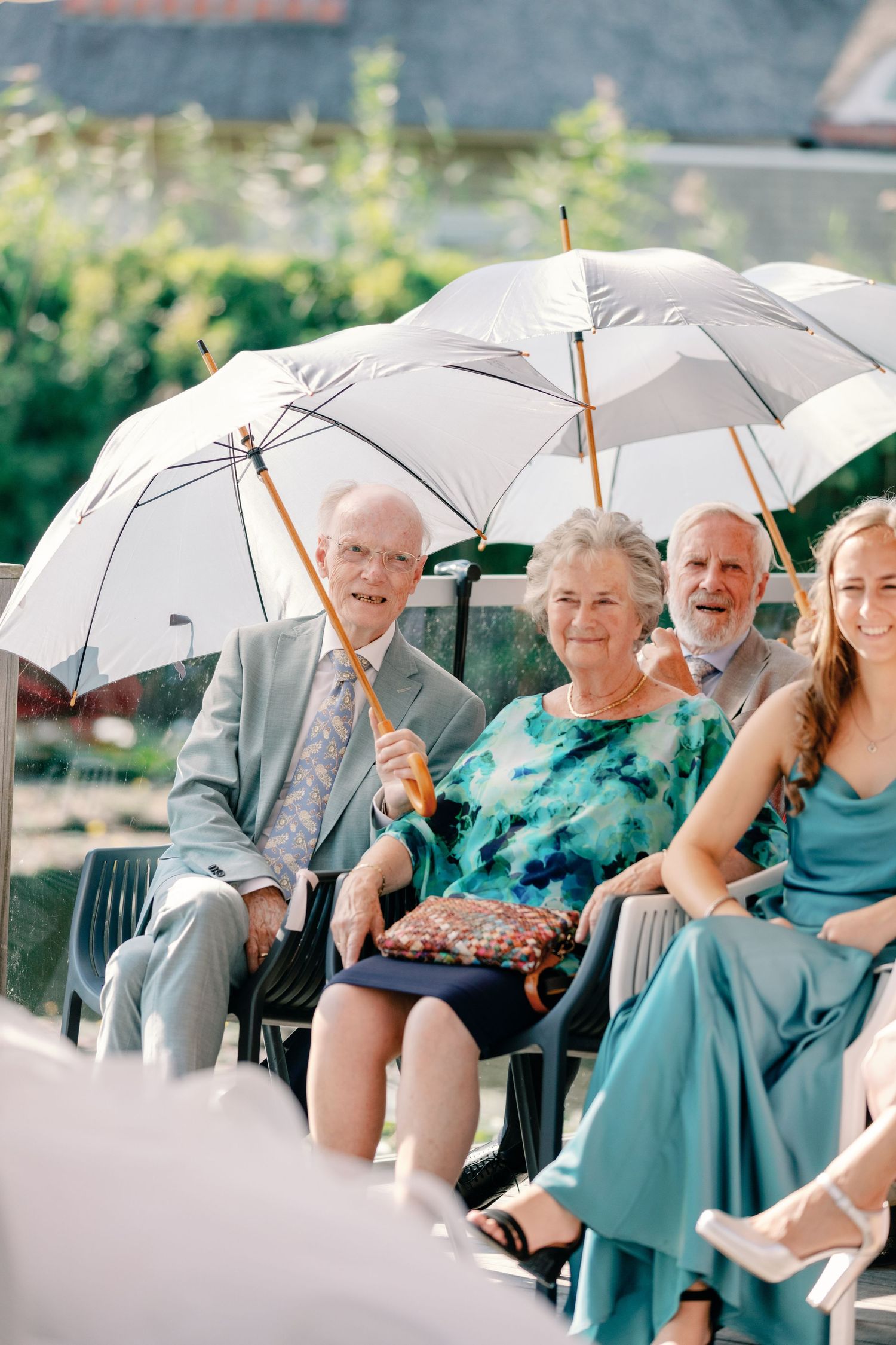 Wedding guests holding white umbrellas smile while seated in outdoor chairs.