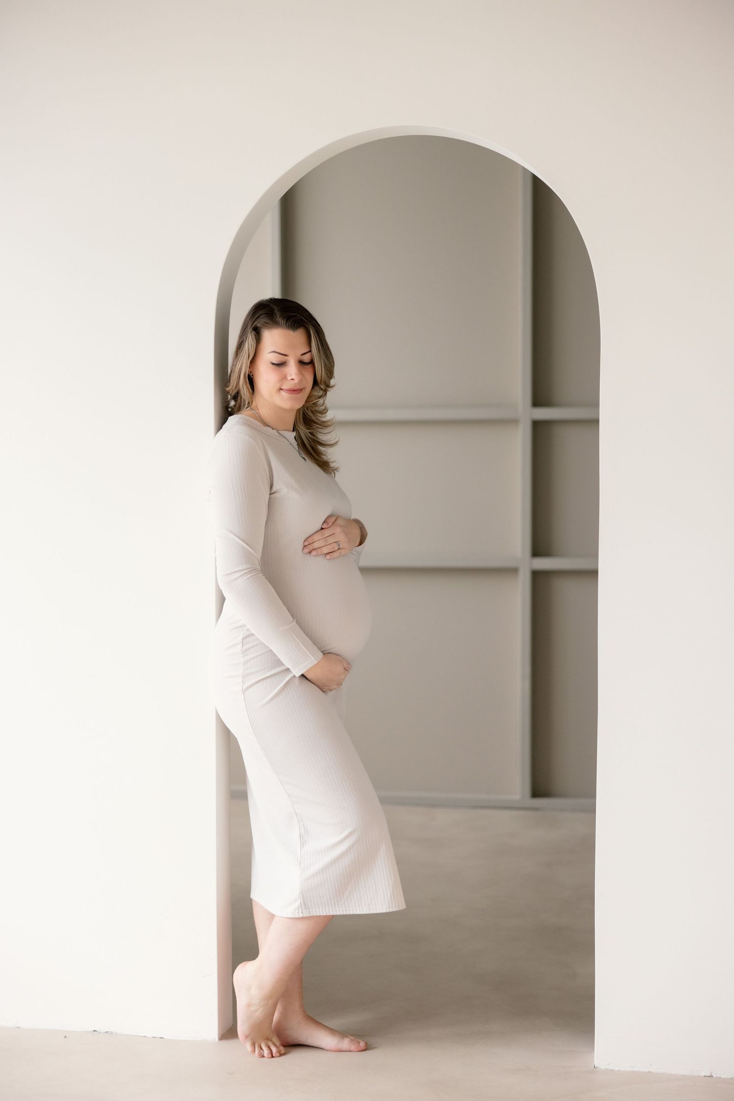 A pregnant woman in a cream-colored dress stands barefoot in an archway against a neutral wall with built-in shelves.