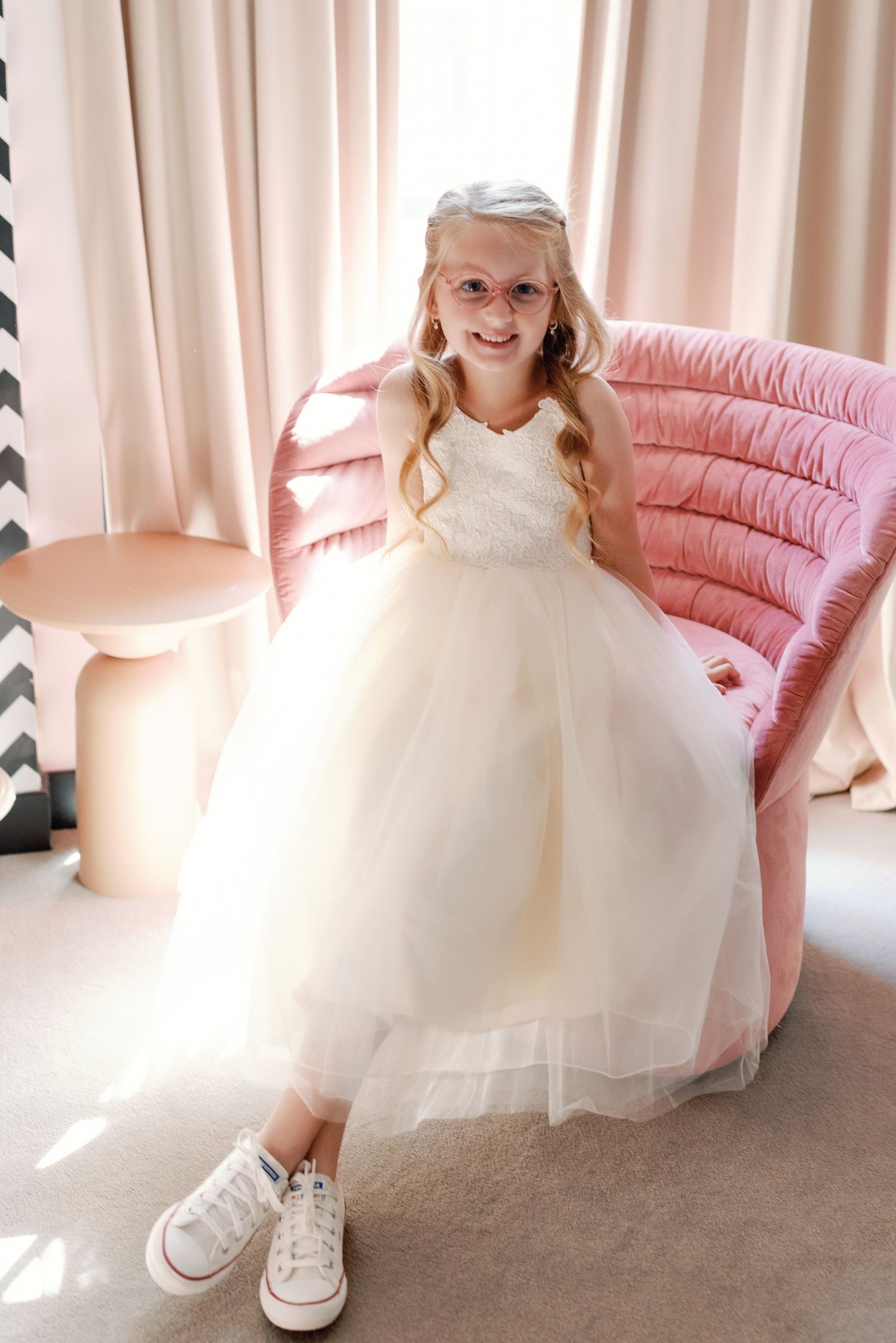 A person in a white tulle dress and sneakers poses on a pink chaise lounge in a sunlit room.
