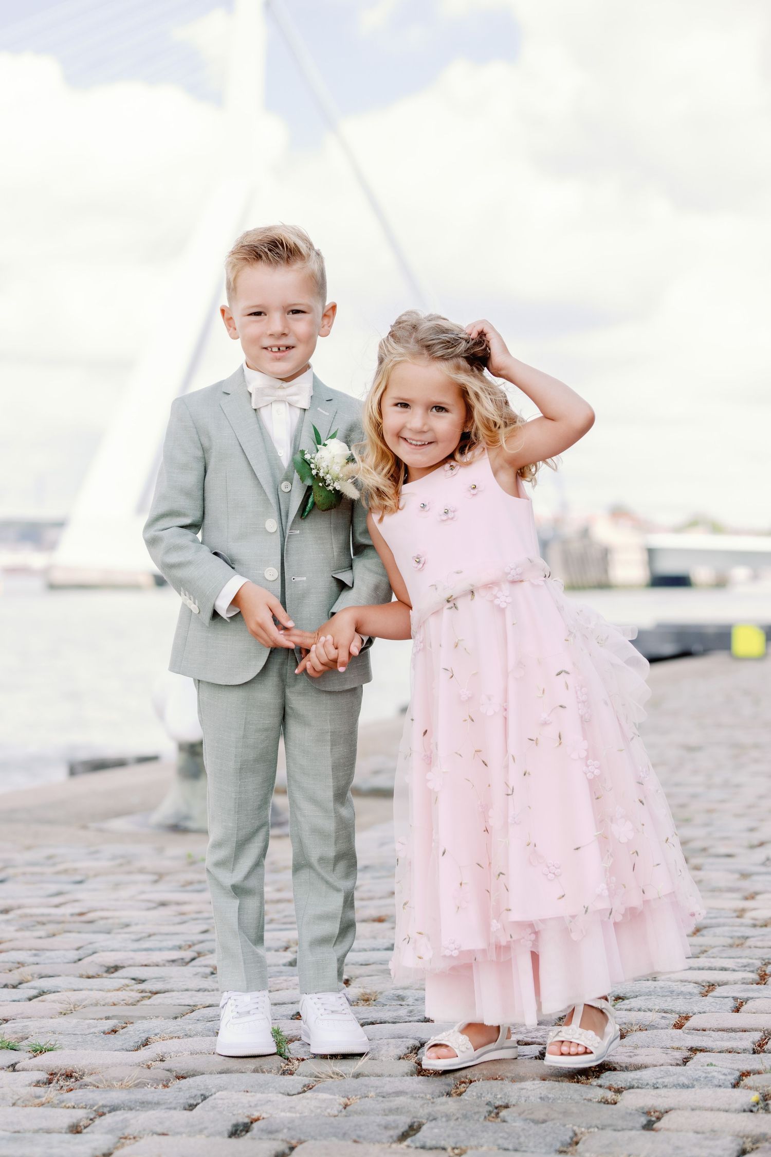 Two children in formal attire, one wearing a gray suit and one in a pink dress, stand together on cobblestone ground.