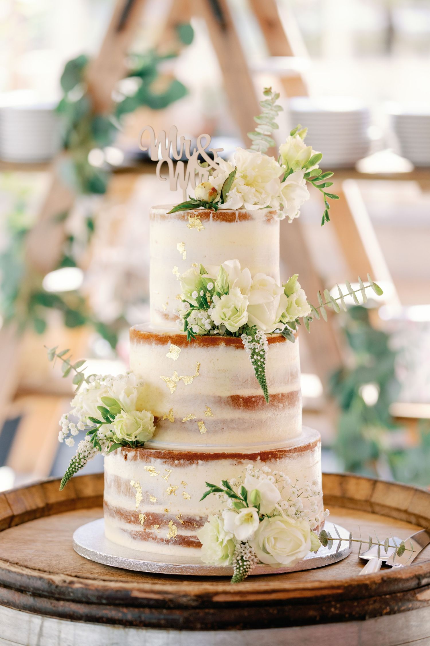 Semi-naked wedding cake with white floral decorations displayed on a rustic wooden barrel platform.