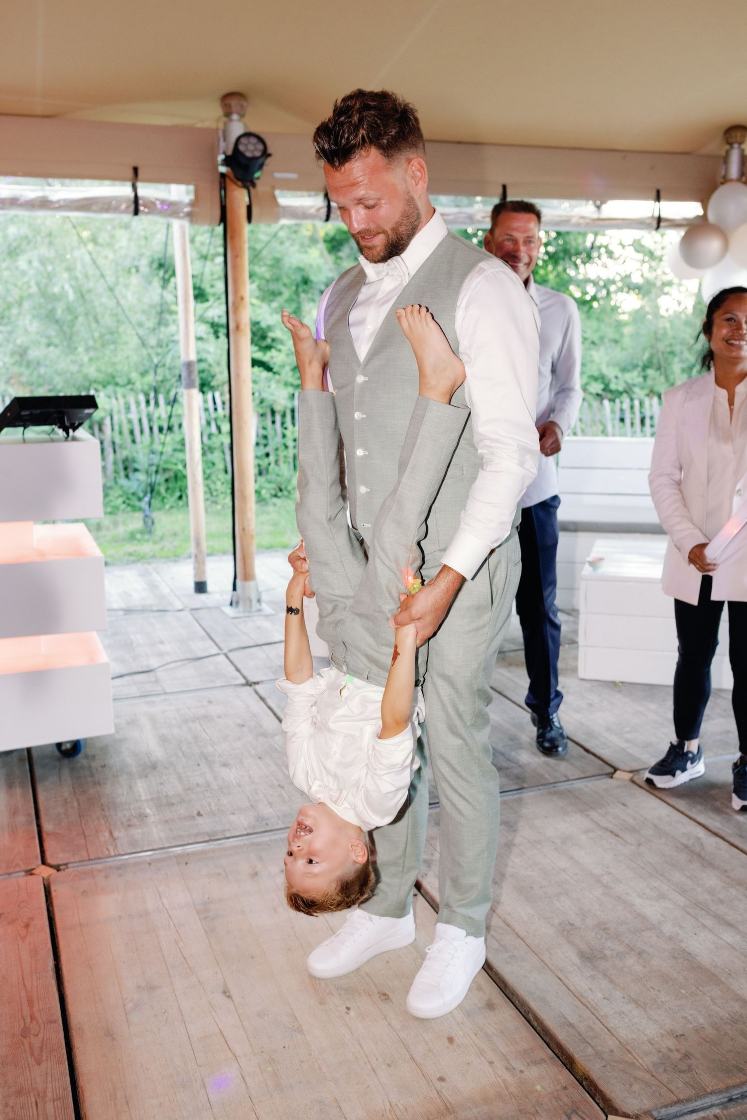A person in a light gray suit holds a small child upside down while dancing at an indoor event space.