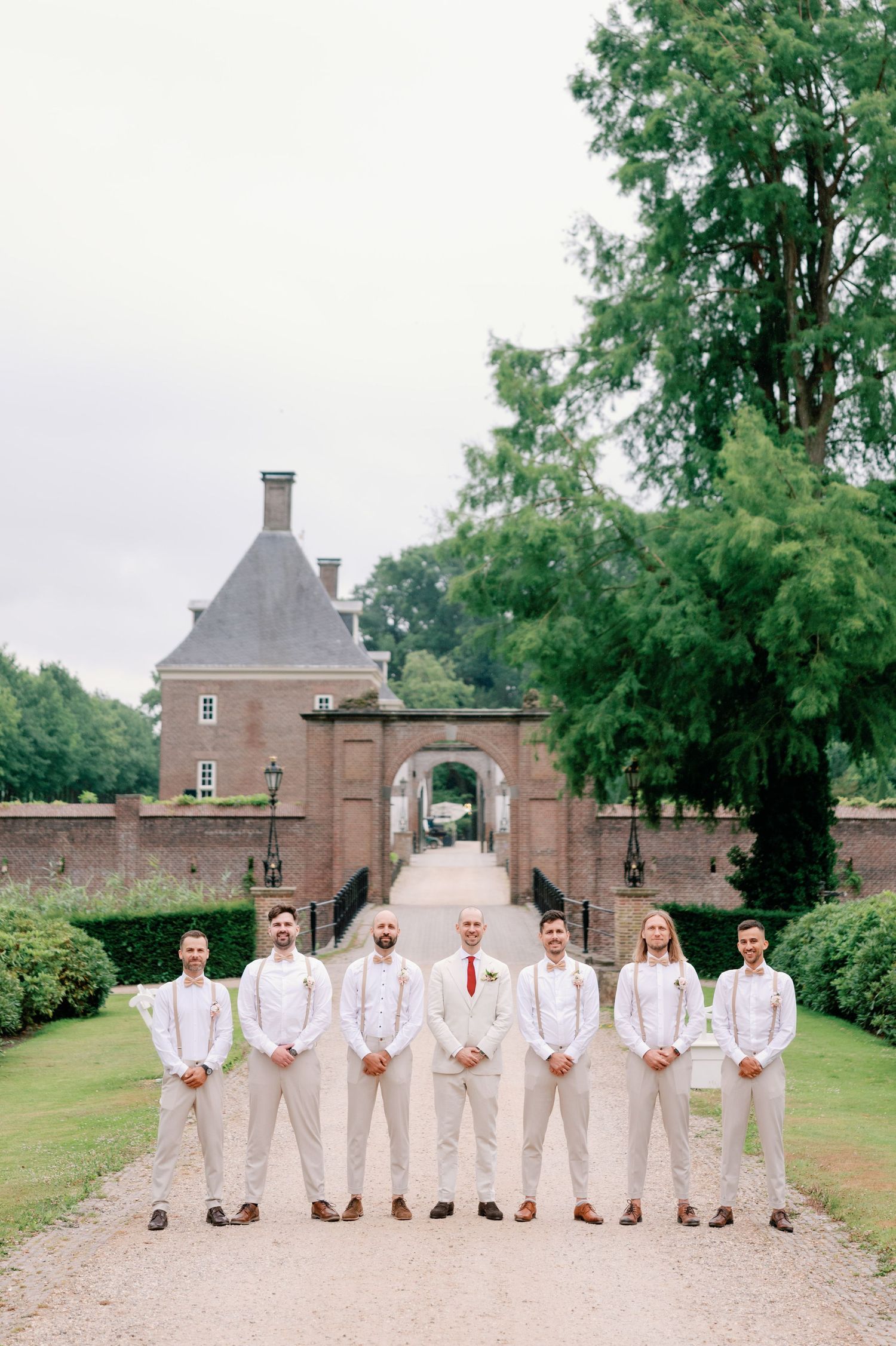 A wedding party in light colored attire poses on a gravel path in front of a historic brick estate entrance.